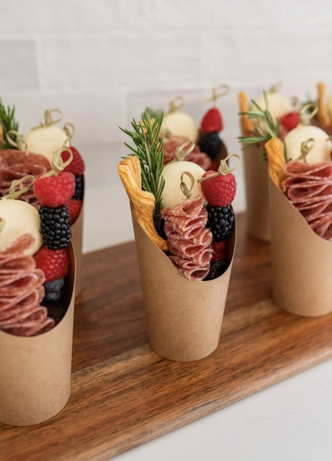 Catered appetizer cones filled with cured meats, cheese balls, raspberries, blackberries, fresh rosemary, and a puff pastry strip on a wooden serving board.