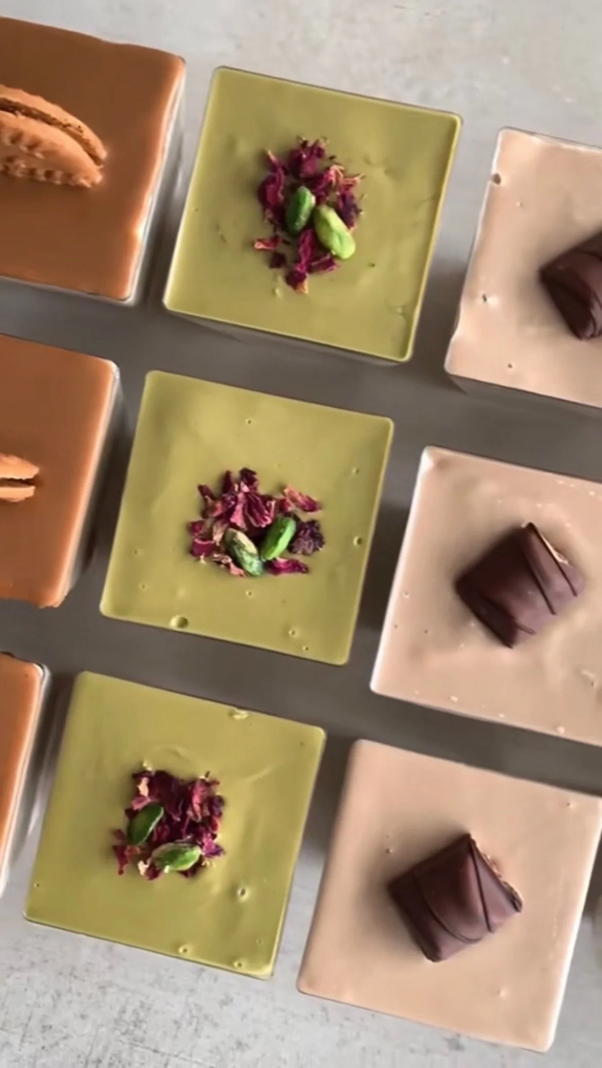 Close-up shot of multiple square cups of different colored chocolates, topped with various garnishes like dried rose petals and pistachios.