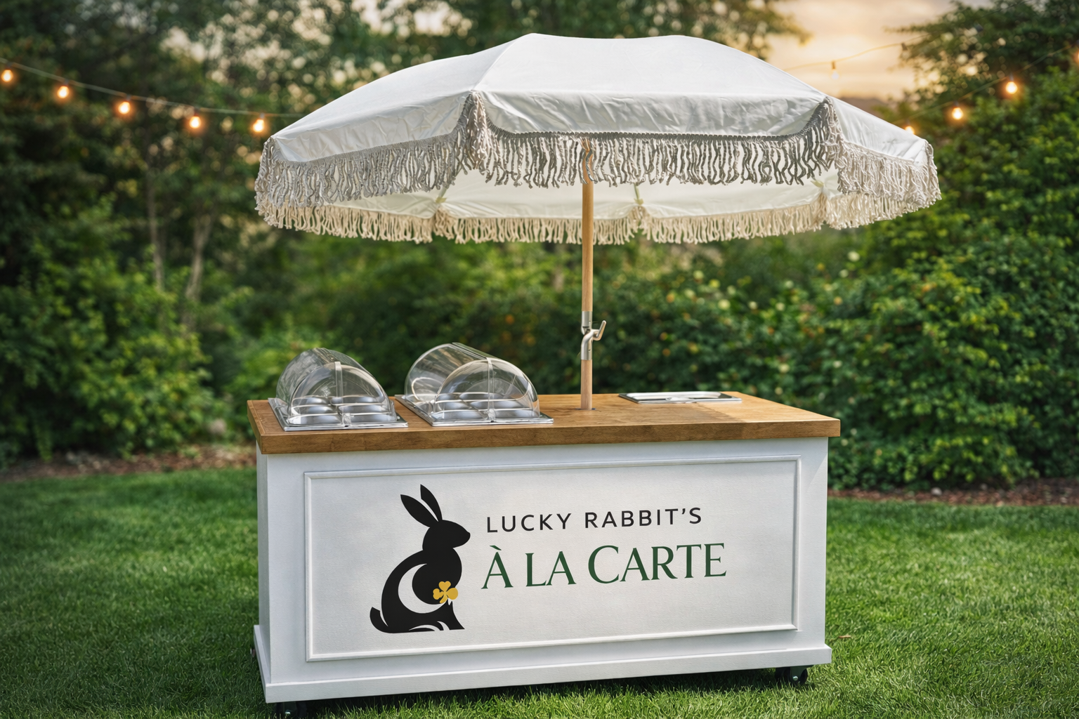 A white outdoor food cart with a wooden top under a large white parasol with fringe, set on a grassy area with trees and string lights in the background. The cart has a sign that reads "Lucky Rabbit's À La Carte" with a black rabbit logo and a small four-leaf clover.