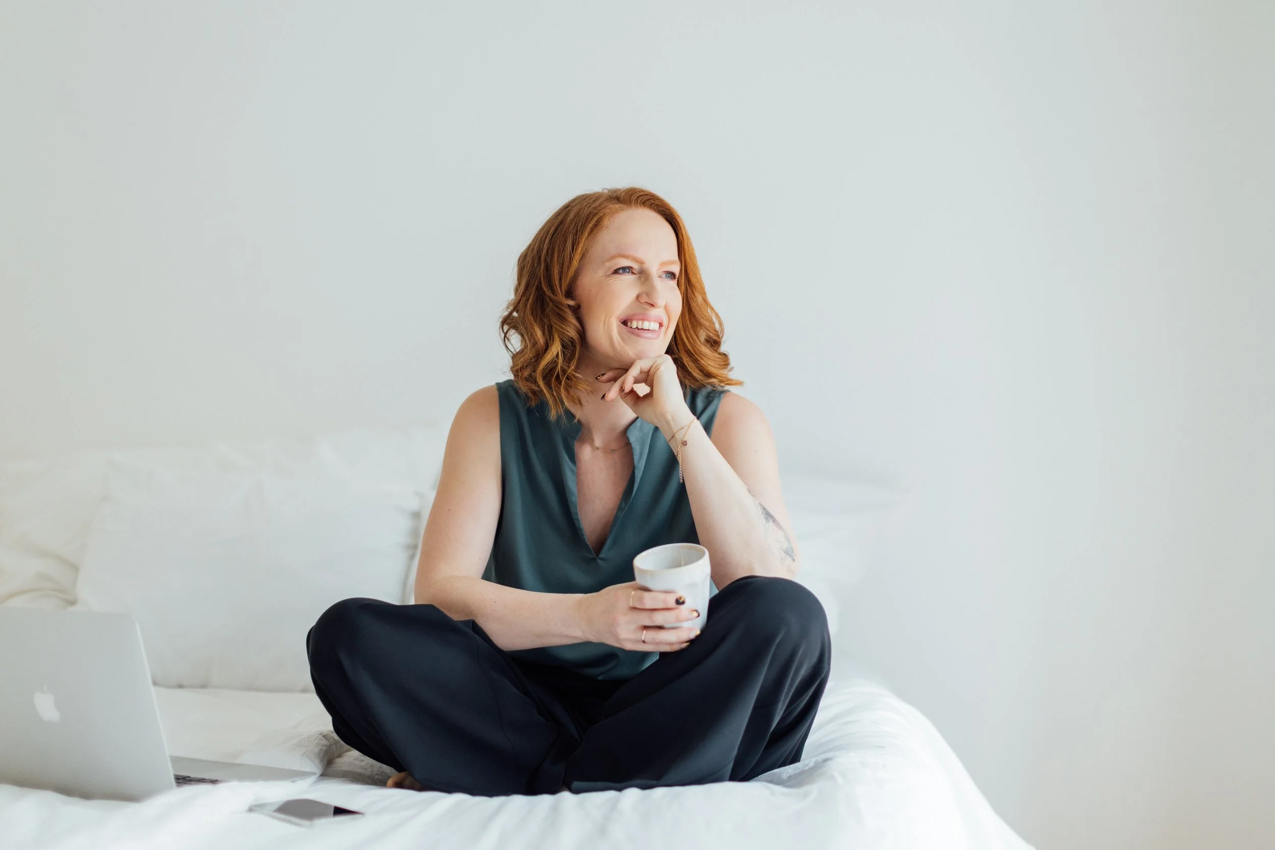 Frau sitzt lächelnd auf einem Bett, hält eine Tasse in der Hand, mit Laptop in der Nähe.
