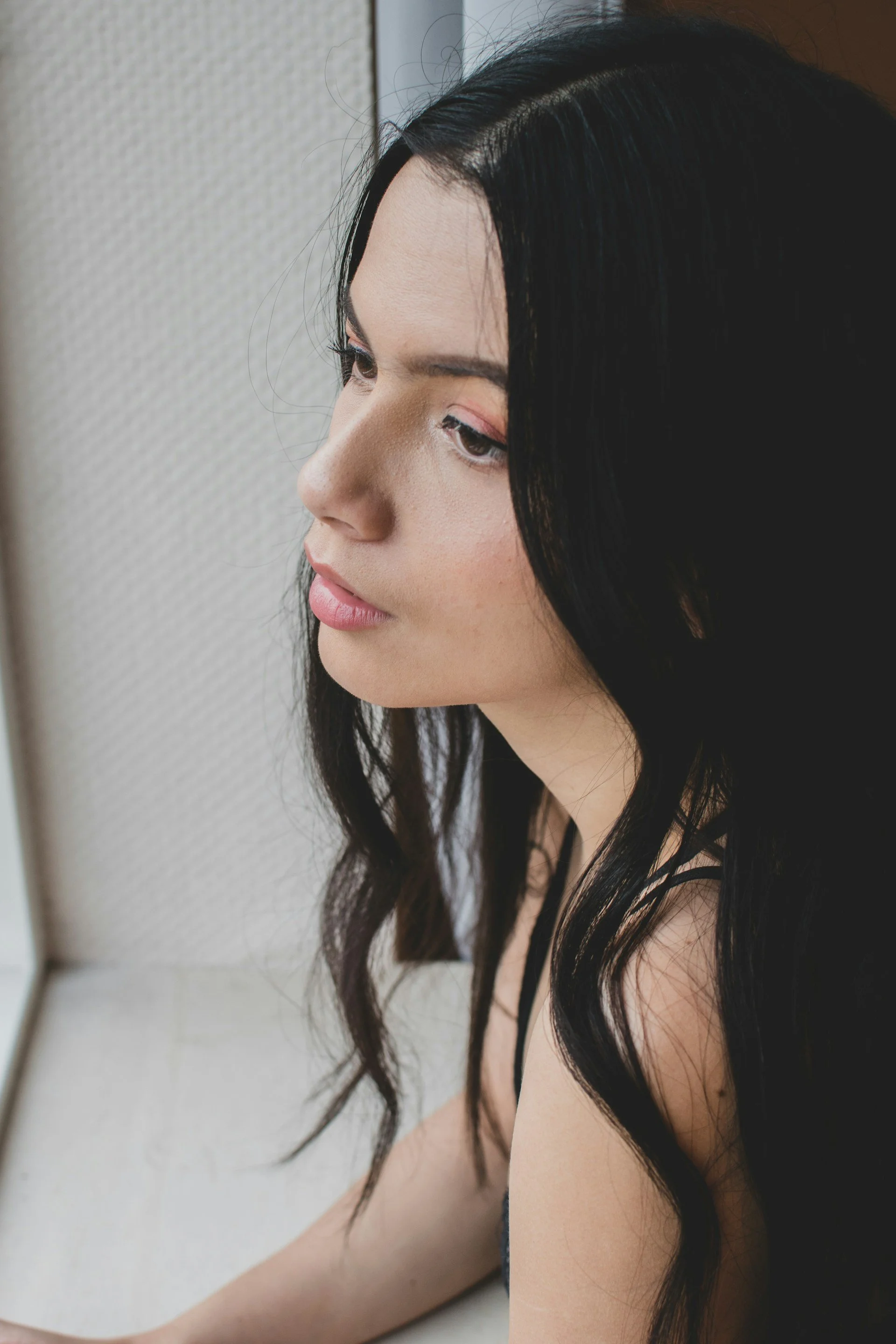 Frau mit langen schwarzen Haaren blickt aus einem Fenster, seitliches Profil, nachdenklicher Ausdruck.