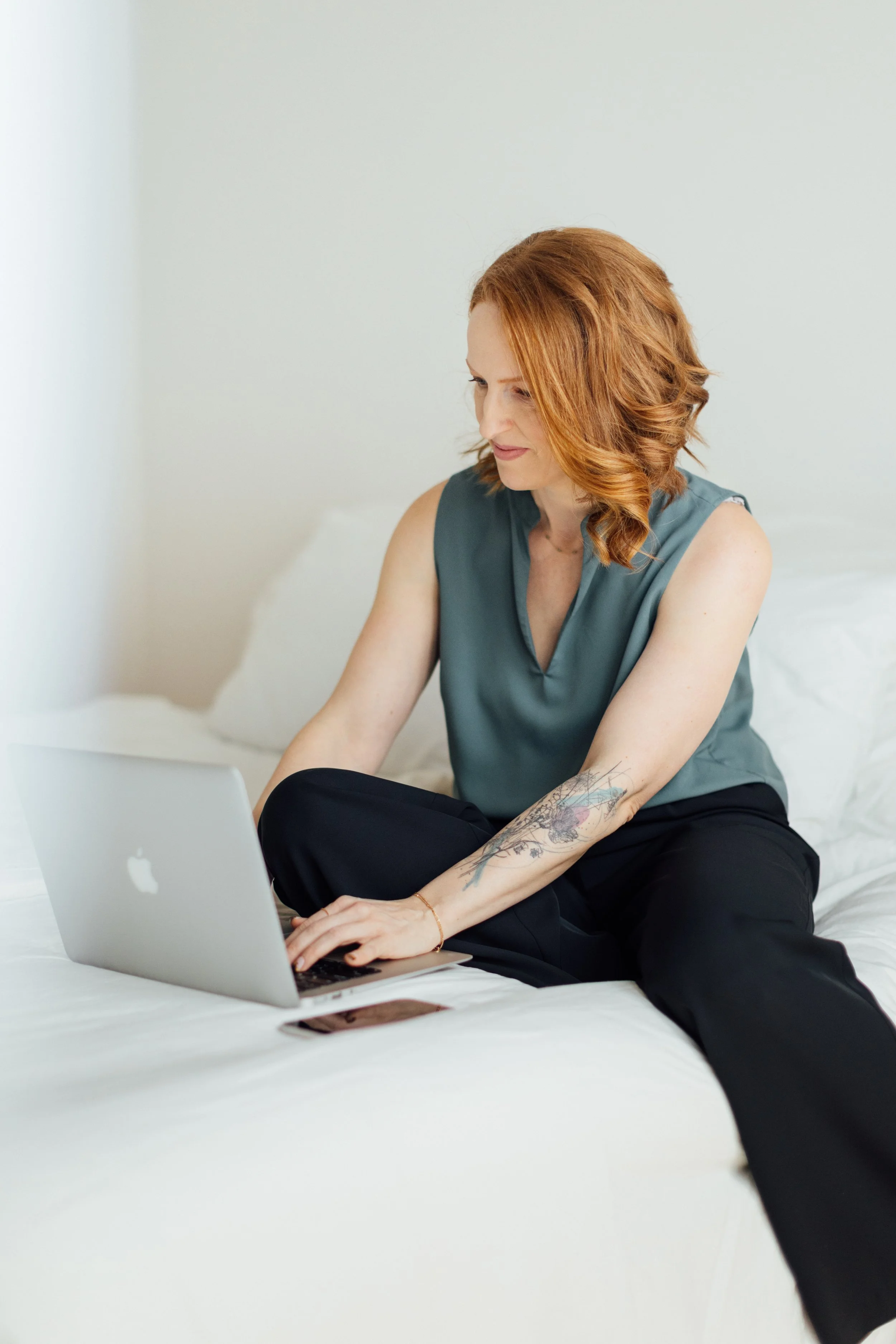 Frau mit roten Haaren und Tätowierung arbeitet auf einem Laptop im Bett.