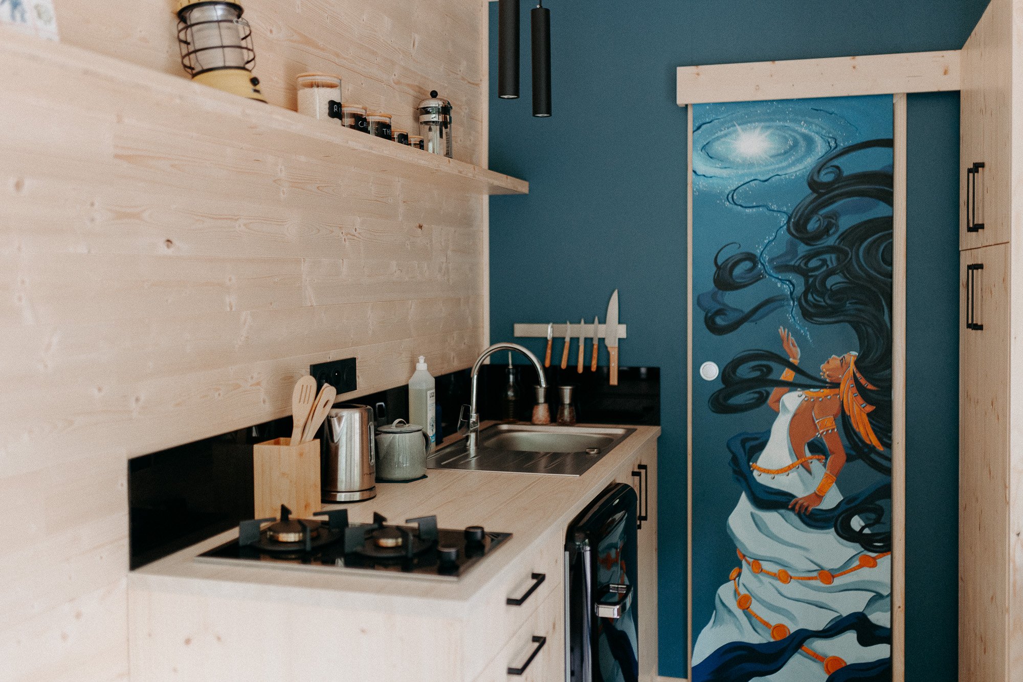 Cuisine avec mur en bois clair, porte décorative avec peinture d'une femme mythologique avec de longues cheveux noirs et un ciel étoilé, accessoires de cuisine, planche à couper, couteaux, lavabo, appareils électroménagers.