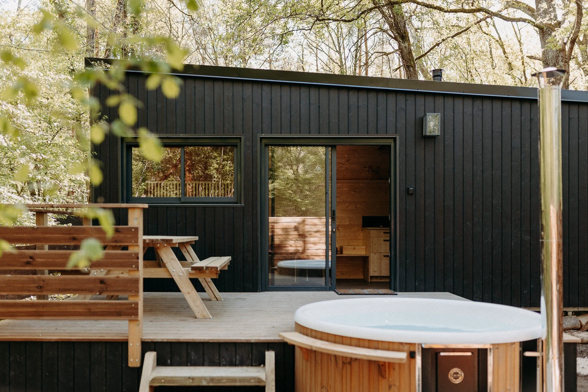 Cabane en bois noire avec terrasse en bois, un jacuzzi en bois, et un mobilier en bois, entourée d'arbres.