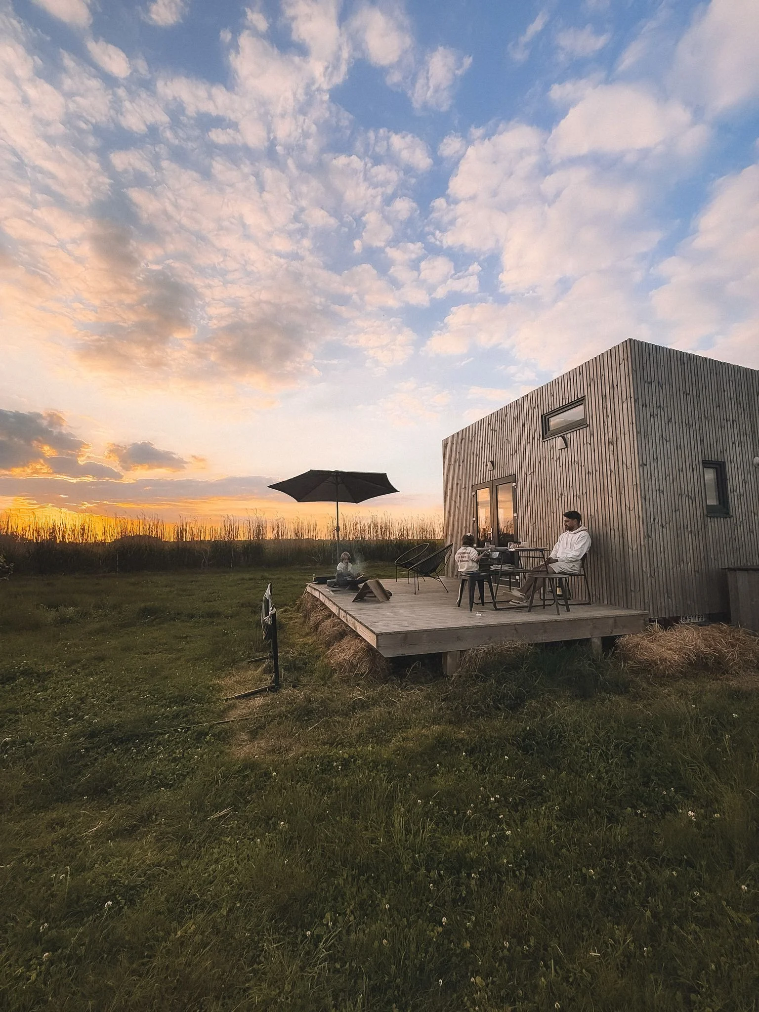 Une maison en bois avec une terrasse en bois, où une famille profite d'un coucher de soleil. La scène se déroule dans un paysage rural avec un ciel partiellement nuageux.