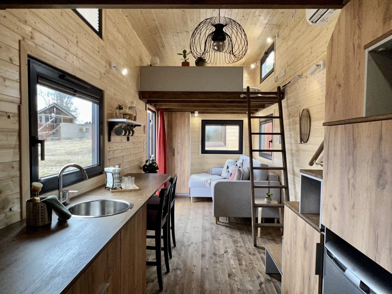 Intérieur d'une petite maison en bois avec cuisine, salon, mezzanine et poutre en bois visible.