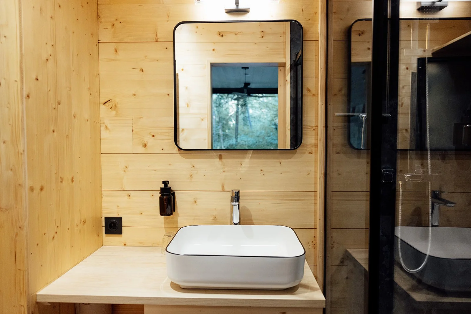 Salle de bain avec un lavabo carré blanc en porcelaine sur un meuble en bois, miroir rond au mur en bois, et cabine de douche en verre avec robinet et pomme de douche.