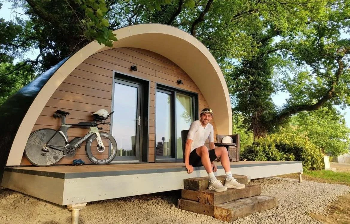 Jeune homme assis sur une petite étape en bois devant une tiny house ronde en bois, entourée d'arbres, avec un vélo posé sur la plateforme