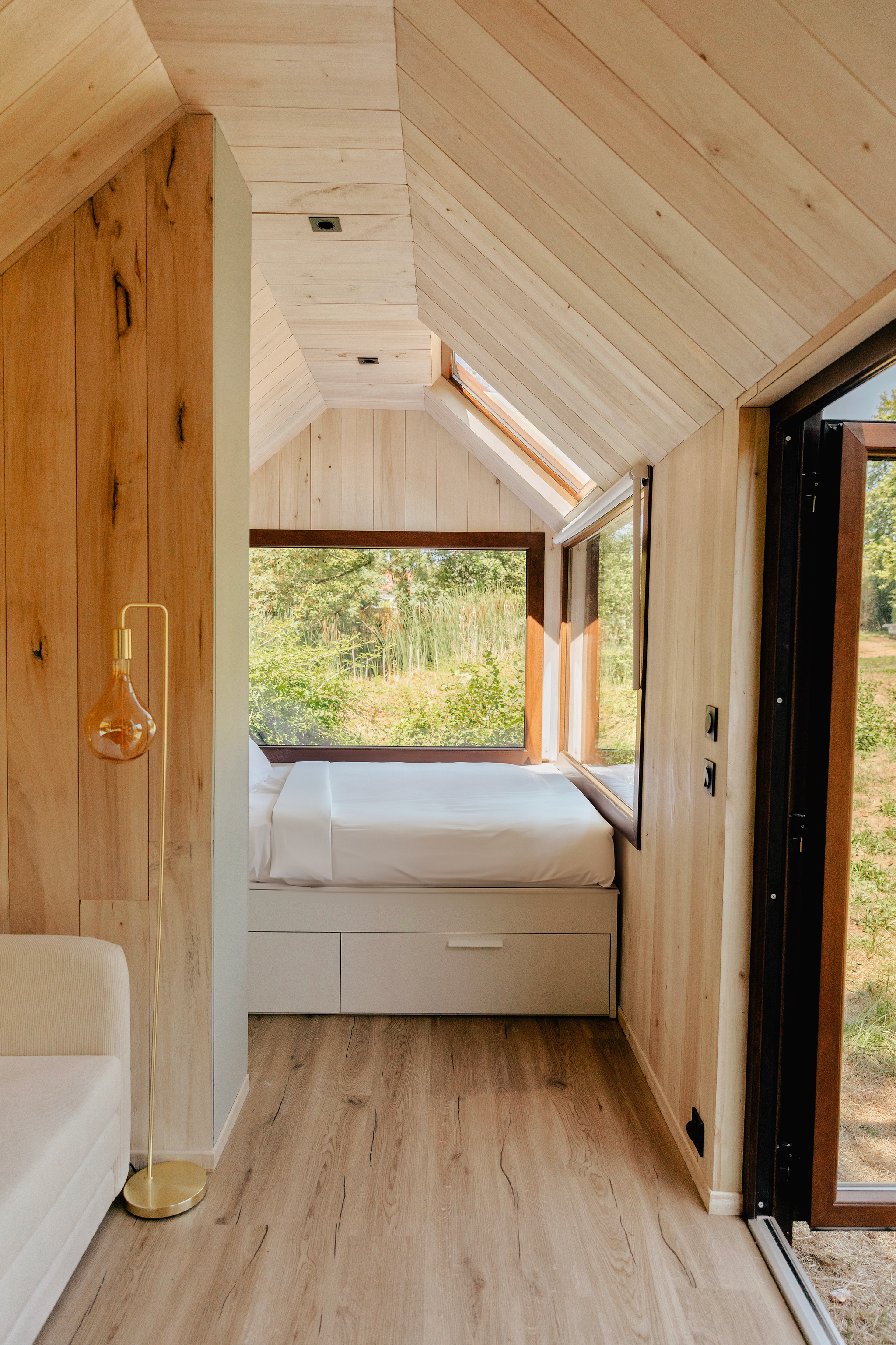Une chambre avec des murs et un plafond en bois clair, une grande fenêtre offrant une vue sur la nature, un lit blanc sous une fenêtre, et un lampadaire à pied doré à côté du canapé beige.