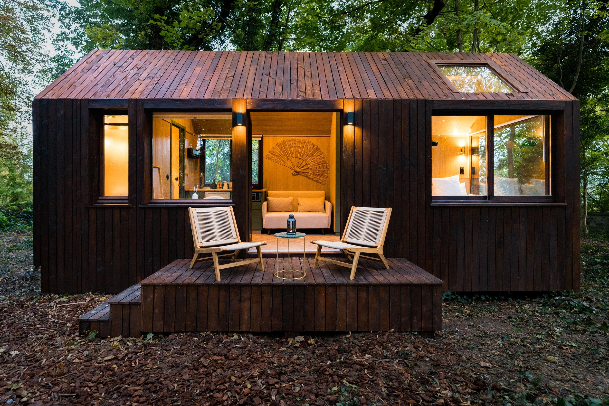 Cabane en bois sombre dans la forêt, avec une petite terrasse extérieure équipée de deux fauteuils en bois et un petit tableau central.