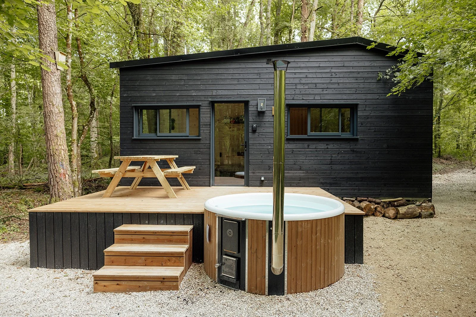 Cabane en bois noir dans la forêt avec une terrasse en bois, un jacuzzi rond en bois, un parasol de soleil fermé, une table de pique-nique en bois, et un chemin de gravier entouré d'arbres