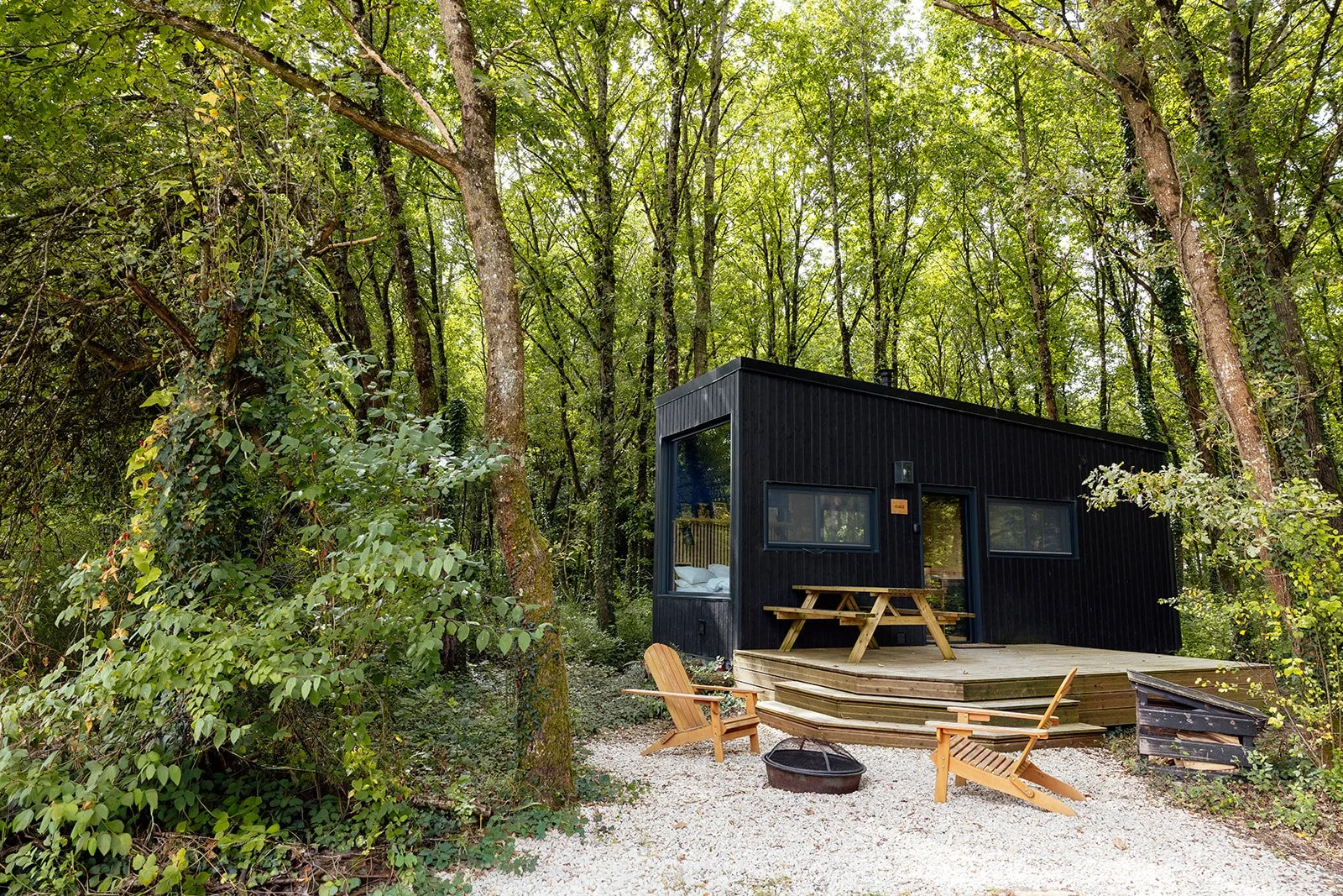 Cabane noire en bois dans une forêt, avec une terrasse en bois, deux chaises en bois, un feu de camp, et un paysage verdoyant.
