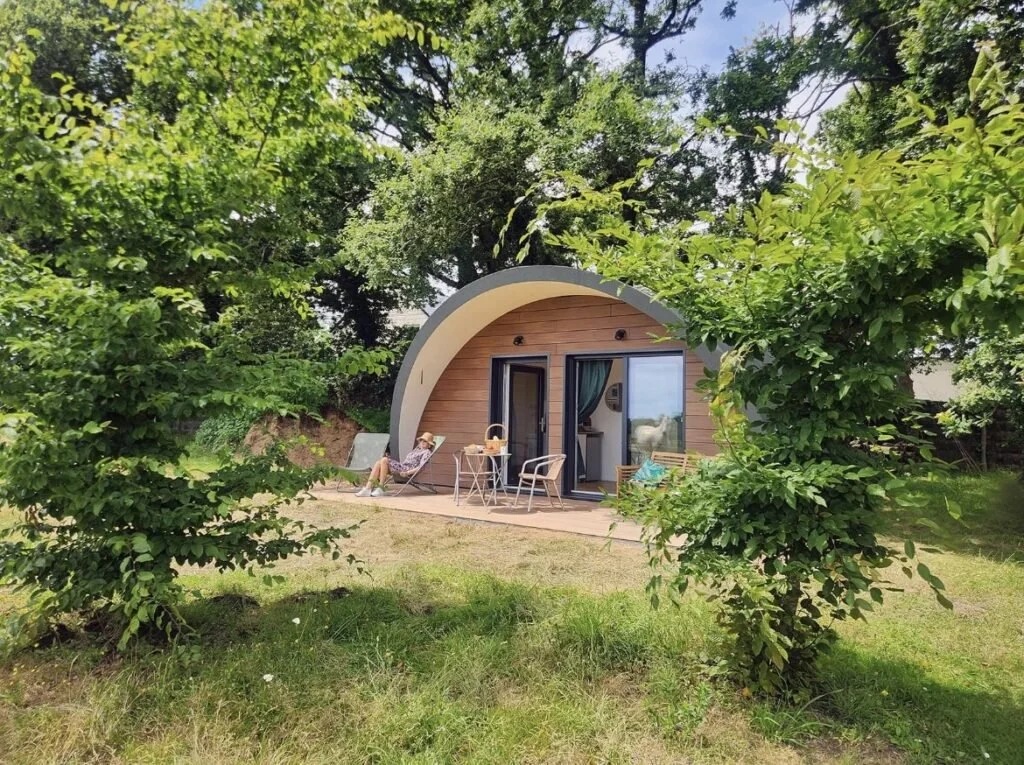 Une petite maison arrondie en bois avec une terrasse, entourée de verdure et d'arbres. Une personne repose sur une chaise longue sur la terrasse.