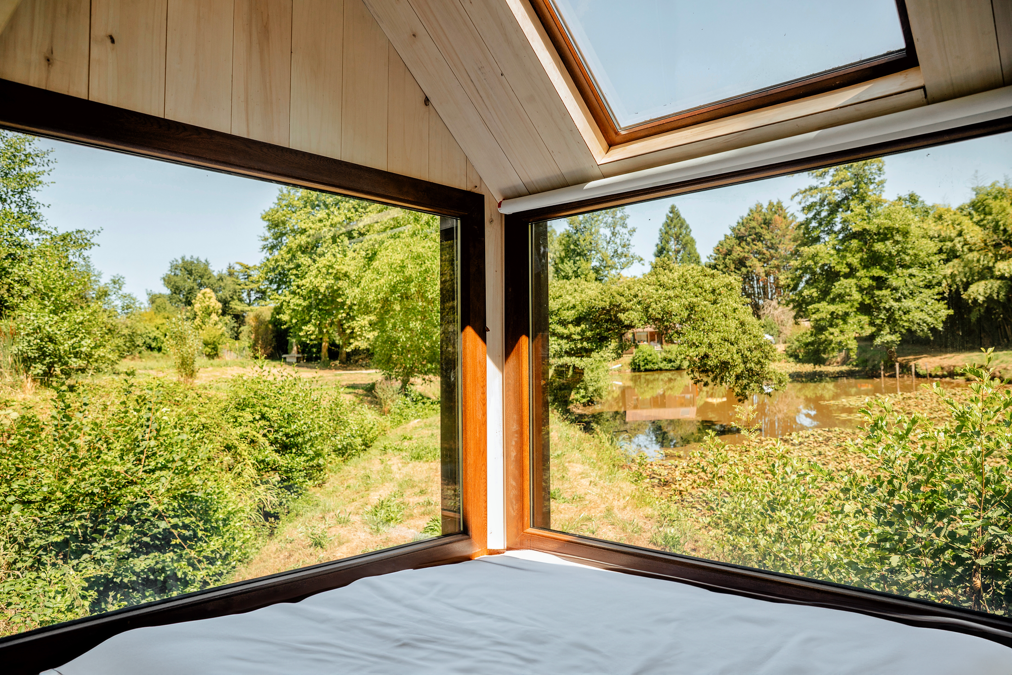 Vue depuis une pièce avec fenêtre panoramique en bois, donnant sur un paysage verdoyant avec des arbres, un lac et un ciel clair.
