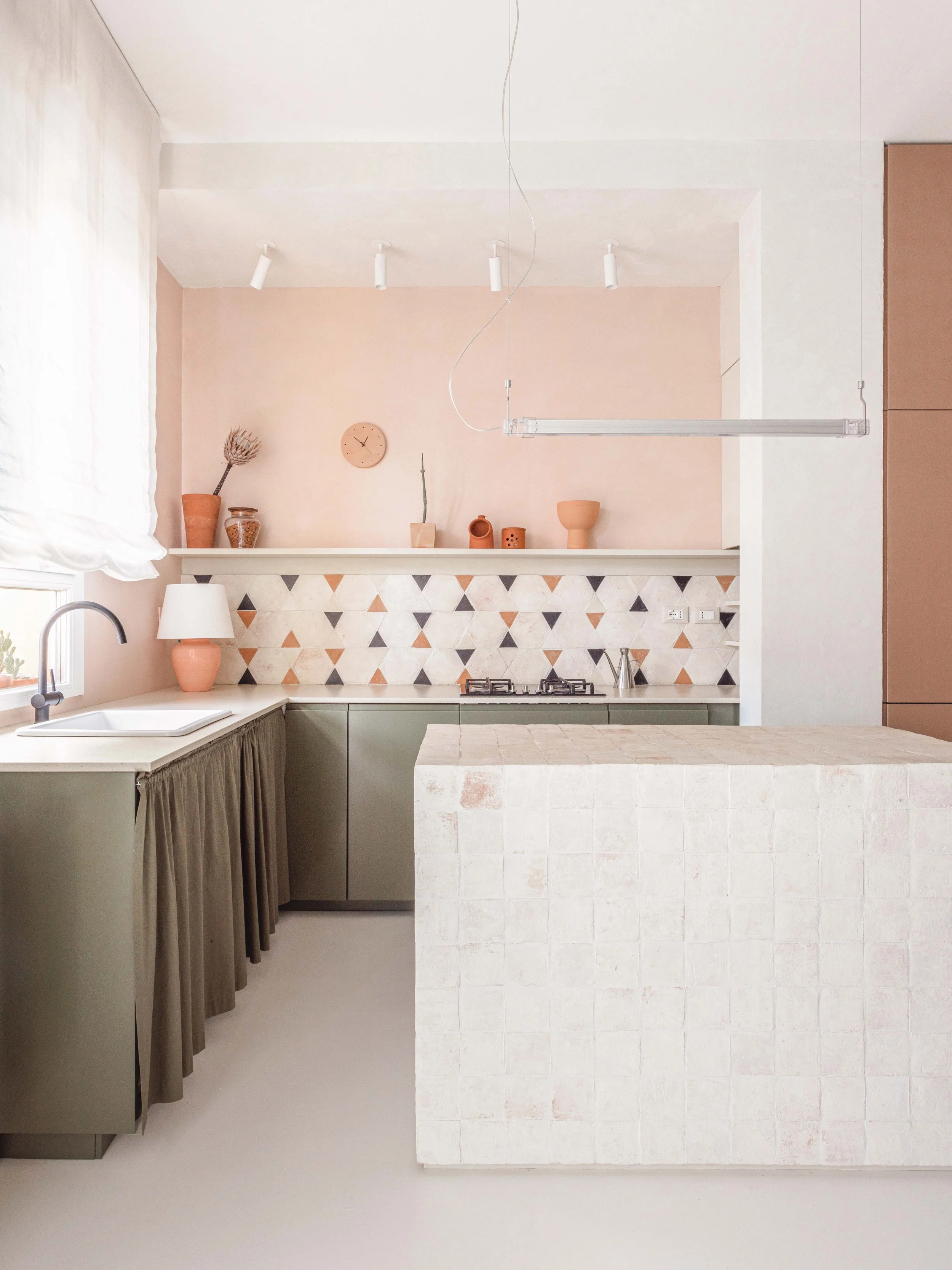 Modern minimalist kitchen with light pink walls, geometric tile backsplash, olive green cabinetry, white countertop, and decorative vases on a ledge.