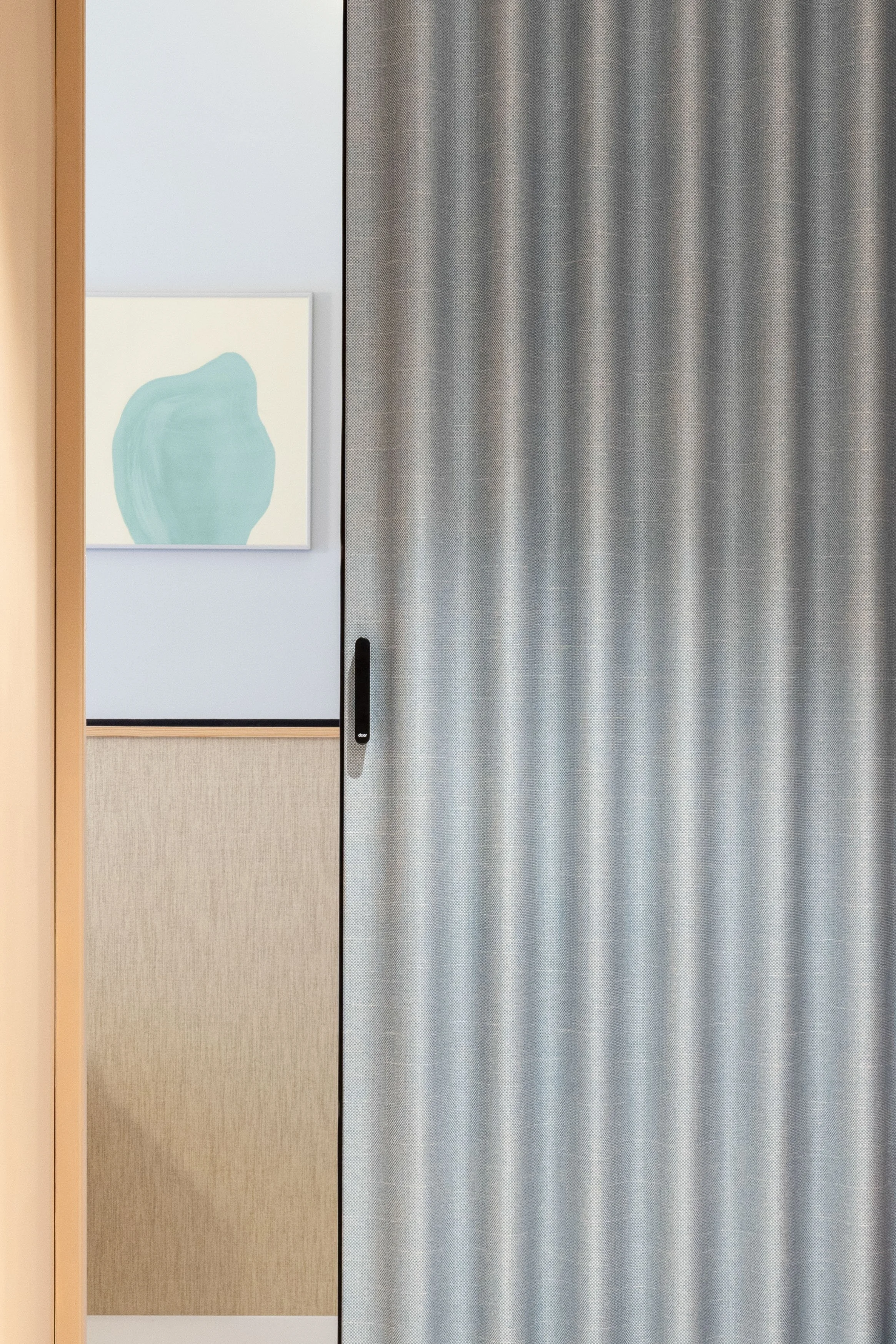 View of a room separated by a sliding grey curtain, with a beige and cream wall, a piece of abstract art, and a wooden door frame.