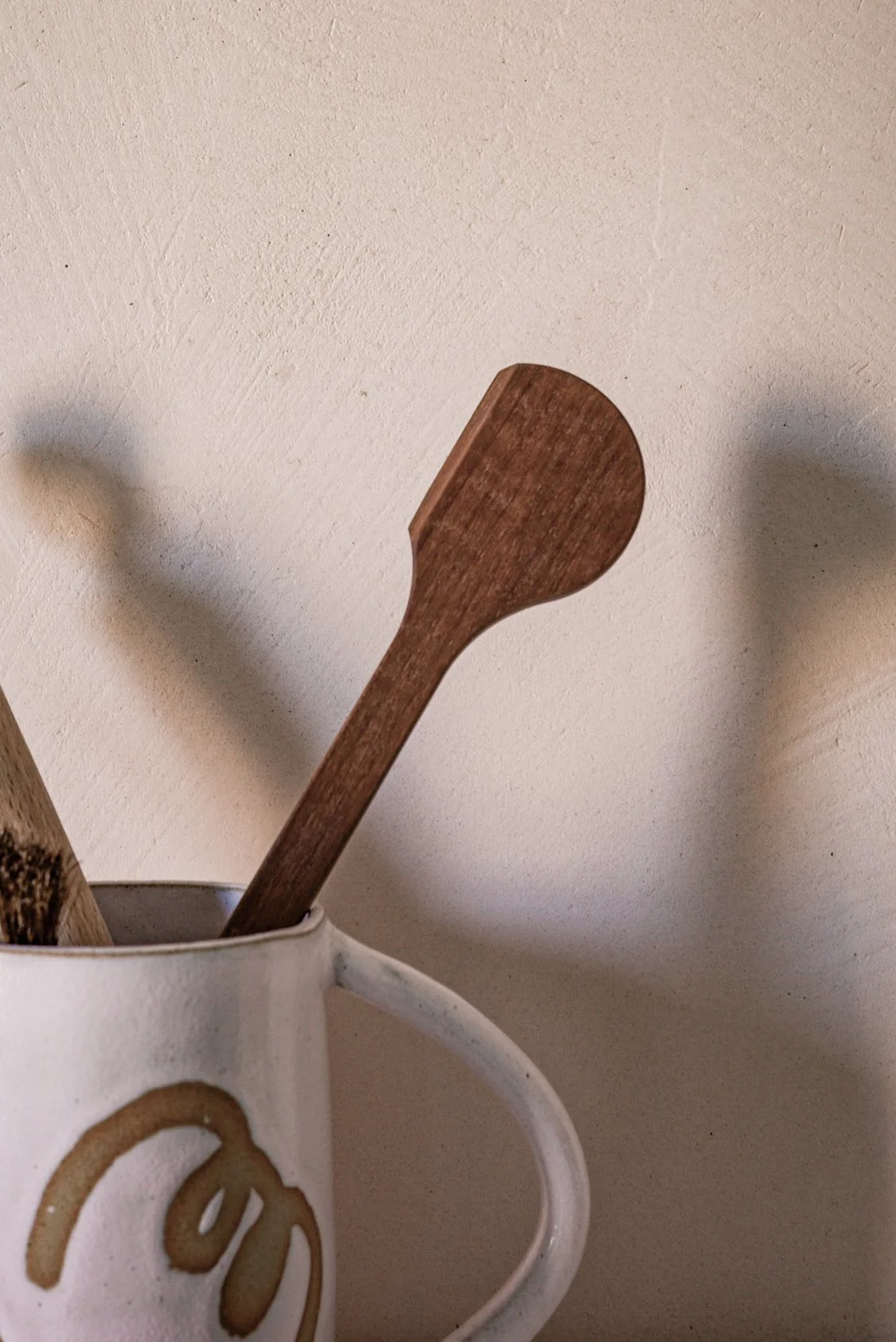 A wooden spoon in a white ceramic mug with a gold-colored handwritten word on it, against a plain beige wall.
