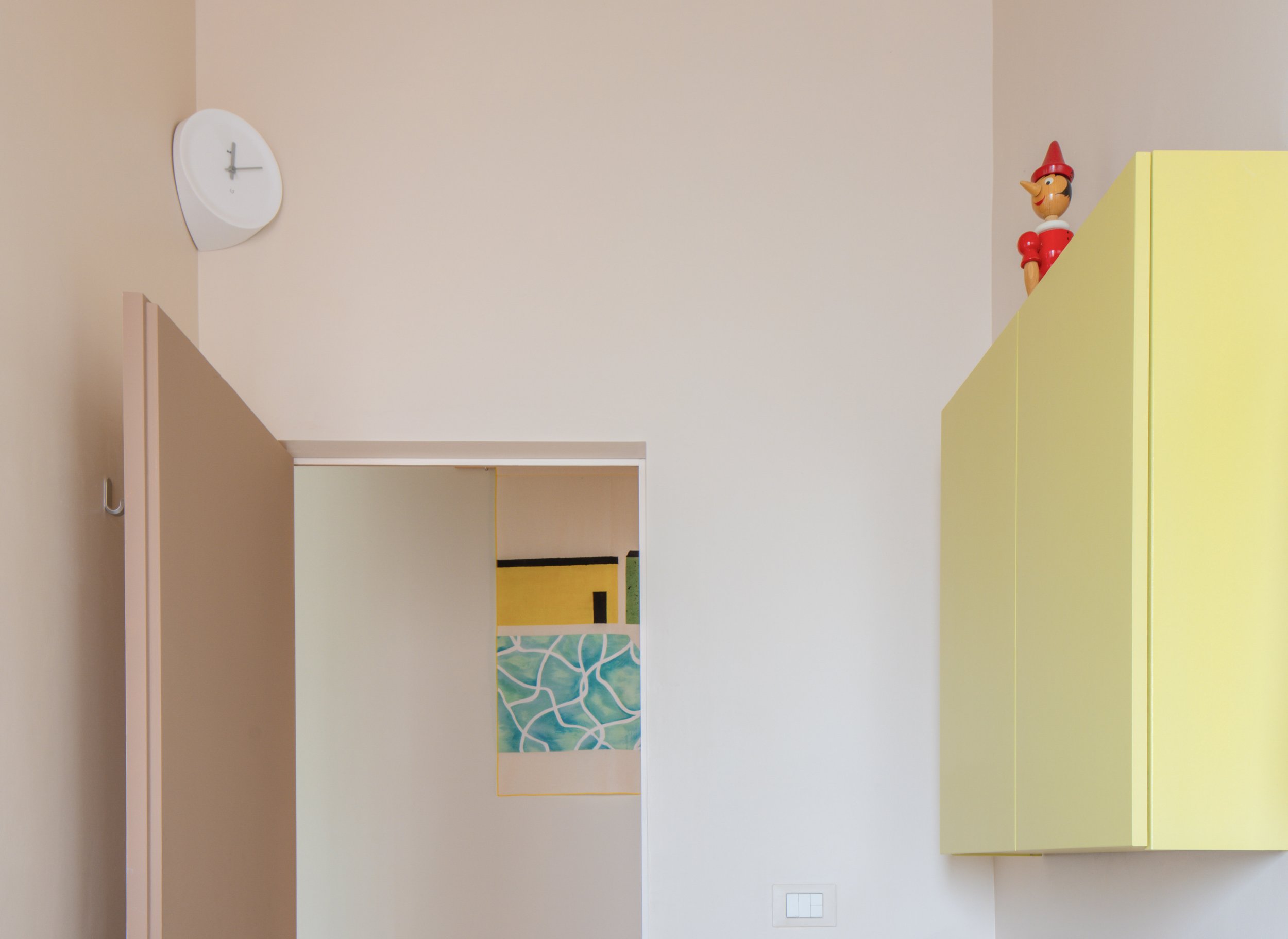 Interior of a room showing a yellow cabinet with a Pinocchio figurine on top, a white wall clock, and a doorway with a painting visible through it.