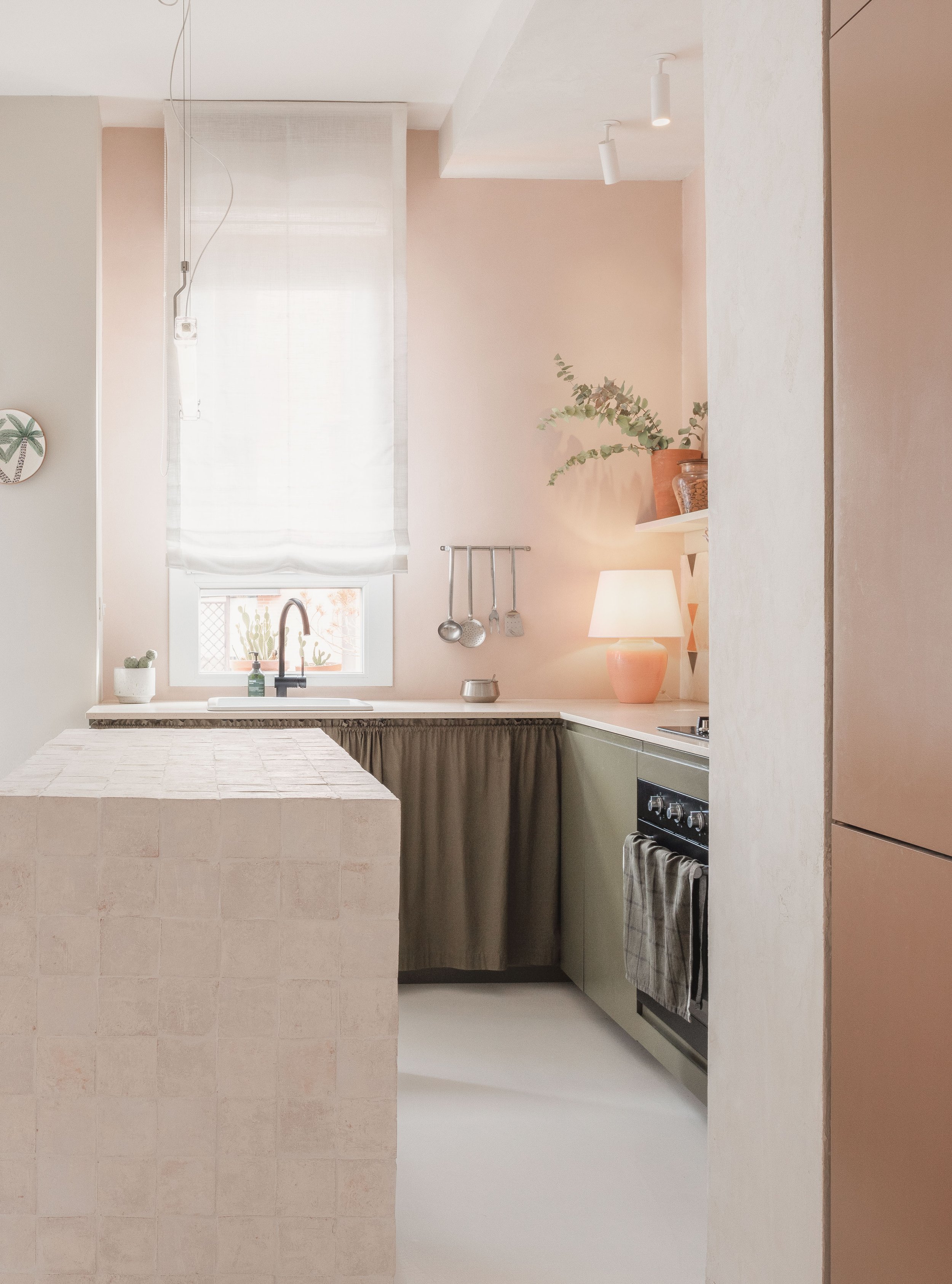 A cozy, minimalist kitchen with beige and light green cabinets, a small window with white blinds, a black faucet, and a black oven. Decor includes a pink table lamp, a large potted plant, and hanging utensils.