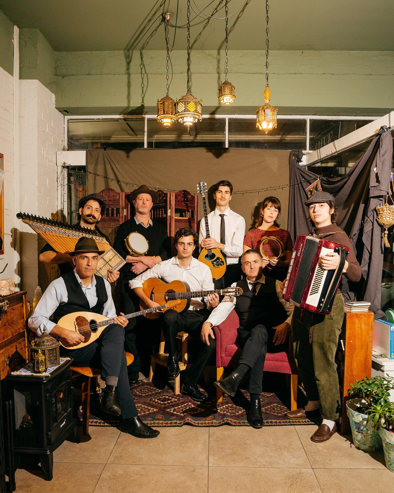 A group of nine musicians, some holding instruments like guitars, tambourine, accordion, and balalaika, pose in a cozy, decorated room with hanging lanterns, a patterned rug, and various accessories. They are posed for a photo, some standing, some sitting, dressed in vintage or classic clothing.
