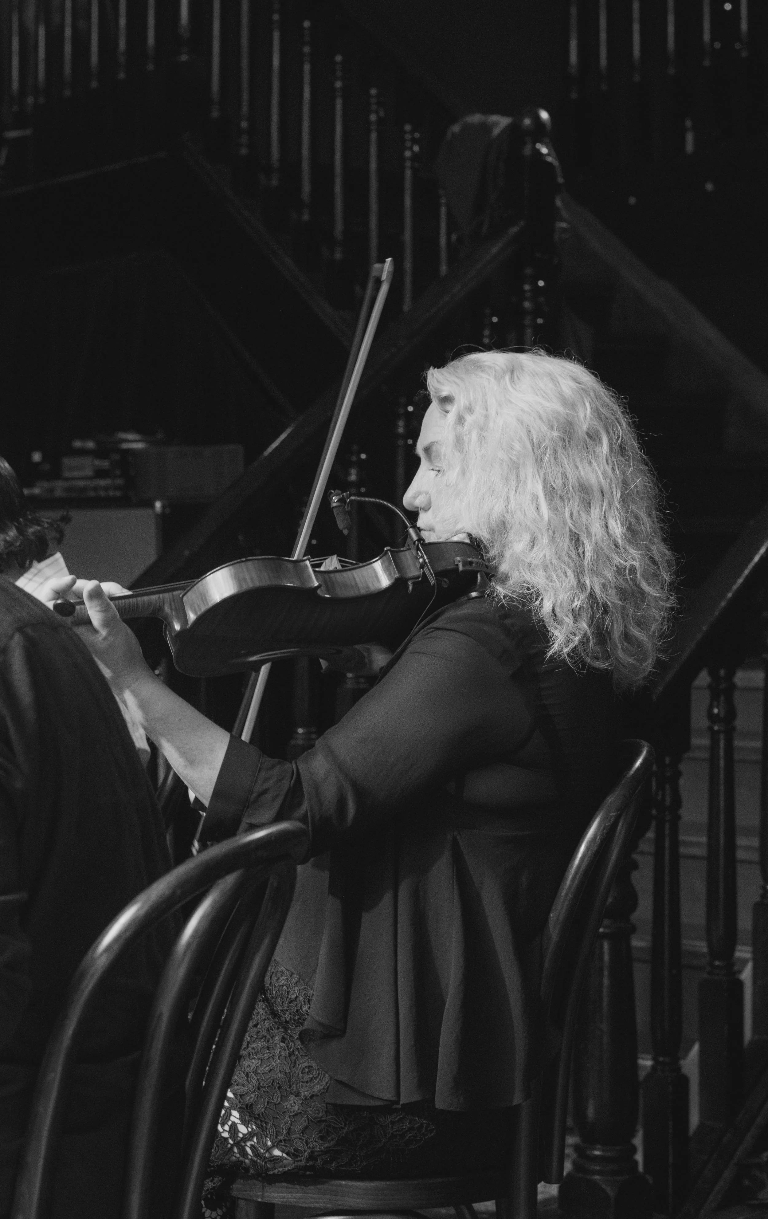 A woman with curly blonde hair playing the violin in a dark room, with a grand piano in the background.