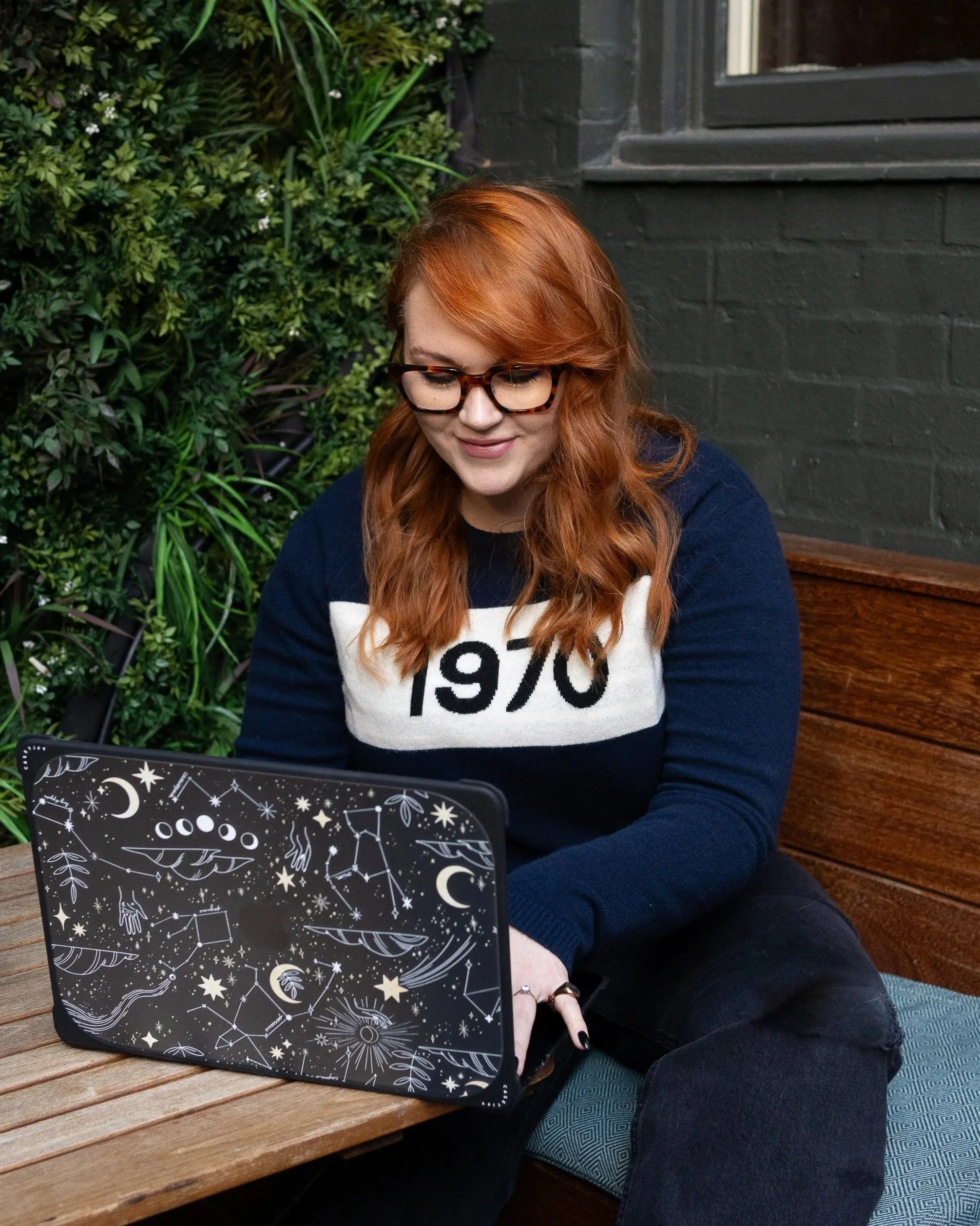 A young woman with red hair and glasses, wearing a navy and white sweater with '1970' written on it, sitting at a wooden table, using a laptop with a celestial-themed design on the cover, in a setting with green plants and a dark gray brick wall.