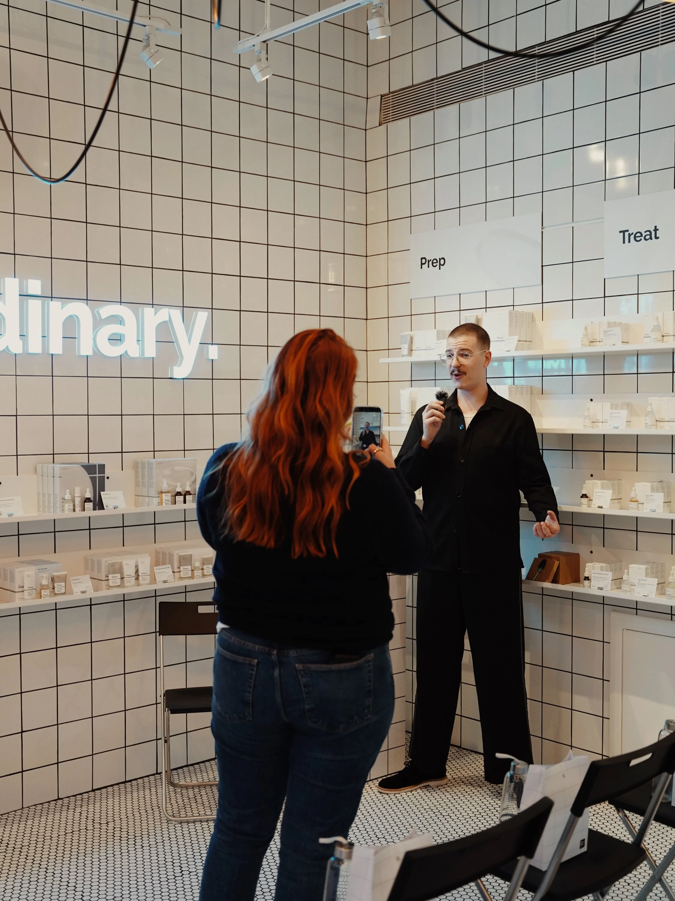A pharmacy with a man and woman engaged in conversation; shelves with medication, signs reading 'Prep' and 'Treat', and a neon sign partly visible that says 'pharmacy.'