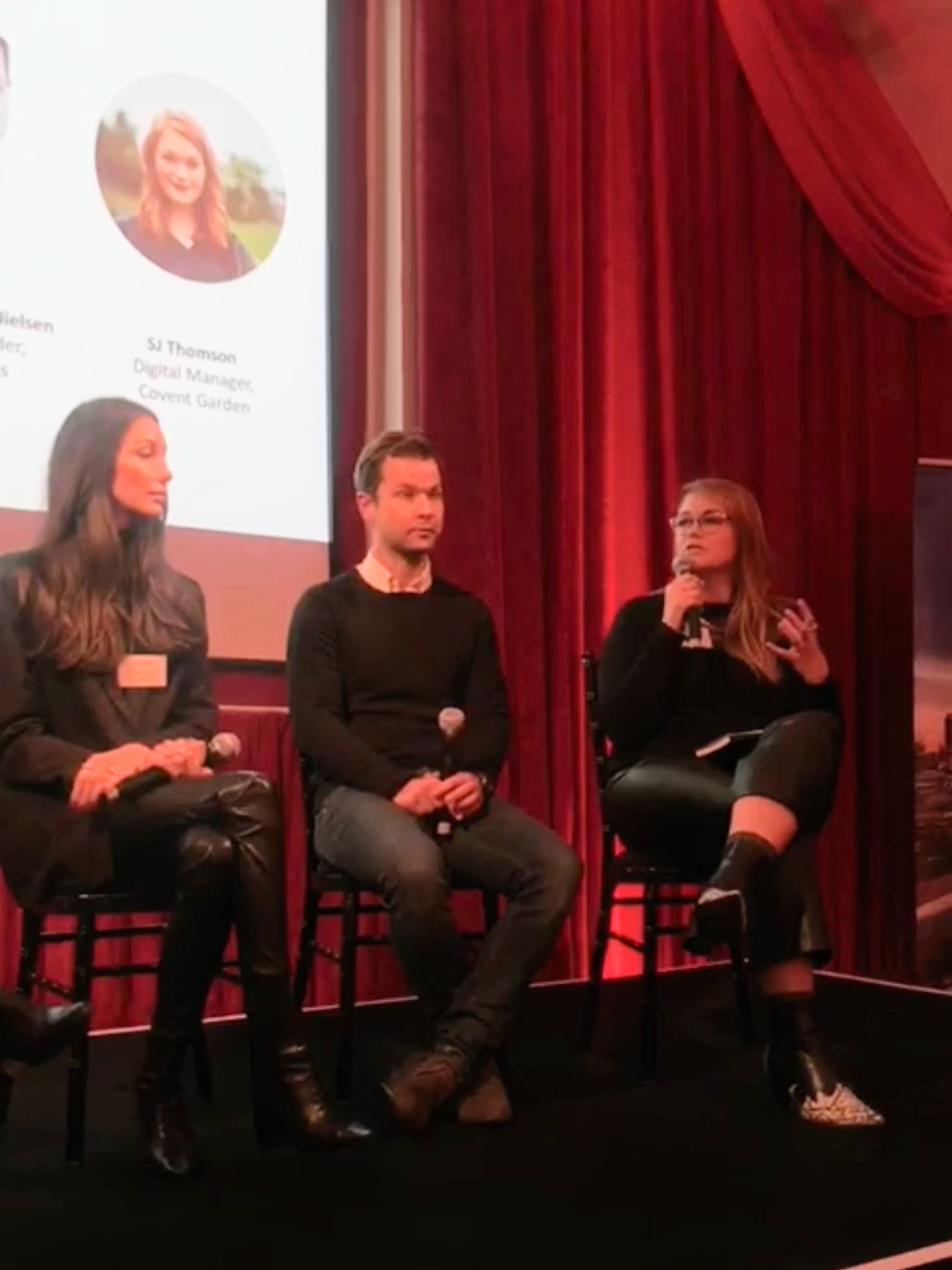 Panel of four individuals seated on stage during a panel discussion, with a woman speaking into a microphone, red curtains in the background, and a large screen displaying a woman's photo and name.