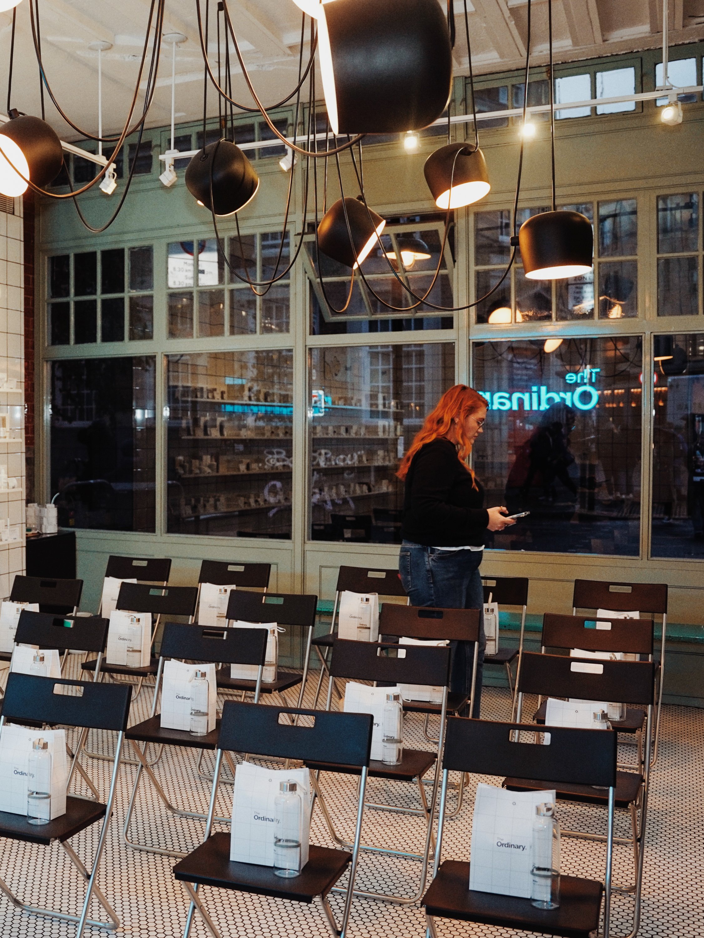 A woman with red hair and glasses standing inside a restaurant, looking at her phone. The restaurant has black chairs, tables with white boxes labeled 'The Ordinary,' water bottles, and a large window revealing a city street outside. The ceiling features modern black pendant lights hanging with visible cords.