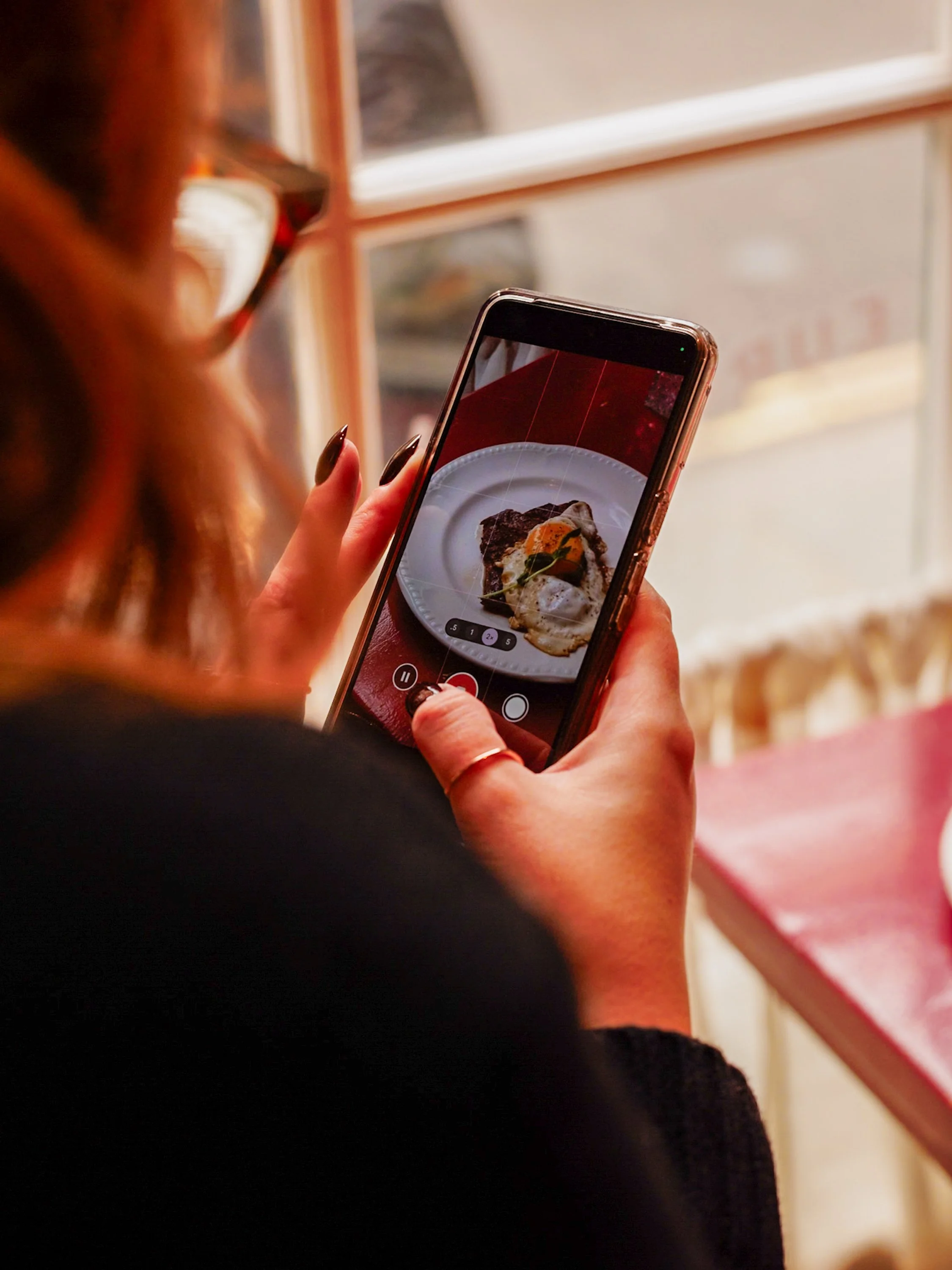 A person holding a smartphone taking a photo of a plate of food, which appears to be a dish featuring eggs, meat, and garnishes.