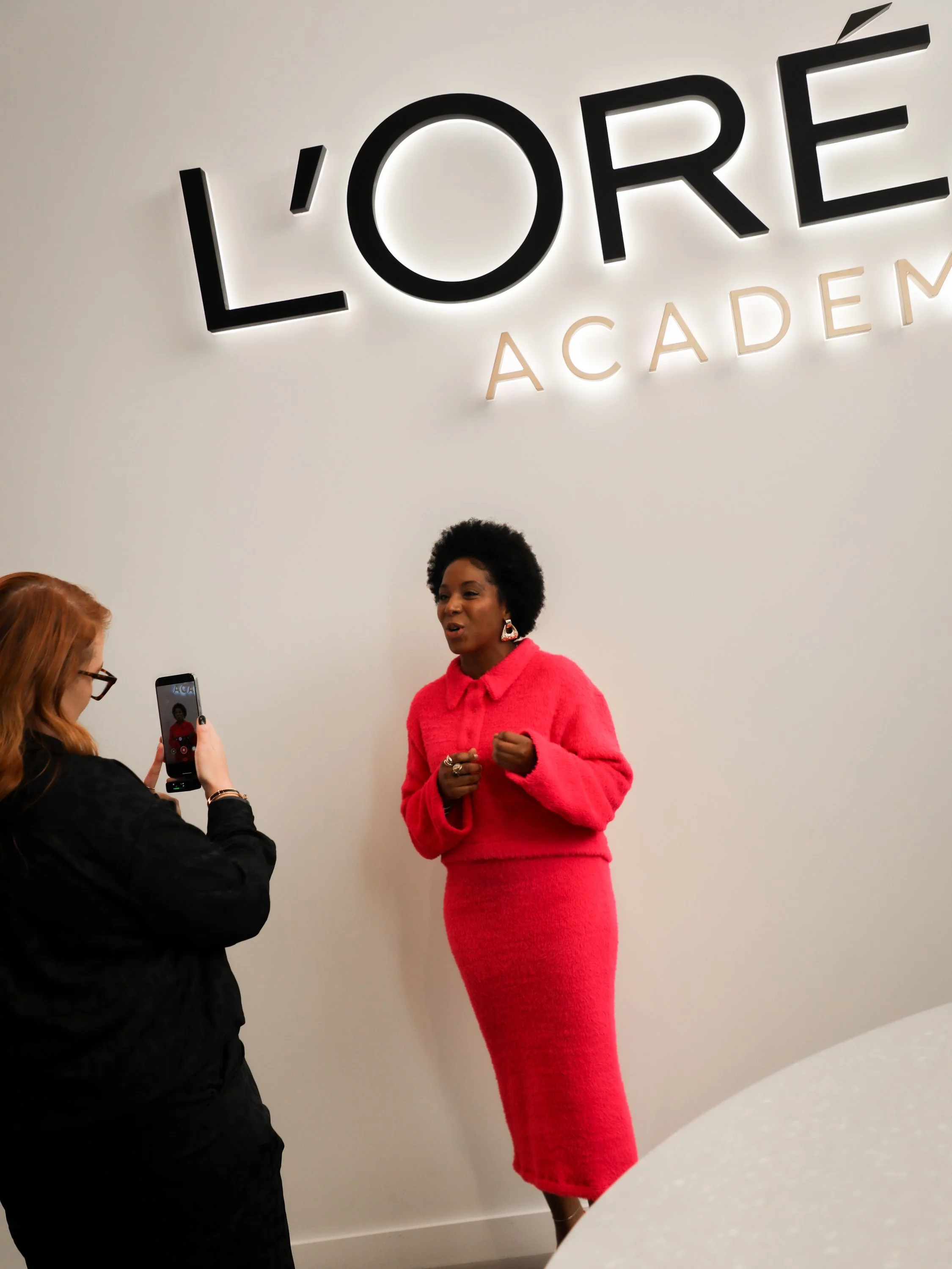 A woman in a bright pink outfit standing in front of a white wall with the words "L'OREE ACADEMY" in large black and beige letters. She appears to be speaking while others are taking pictures of her.