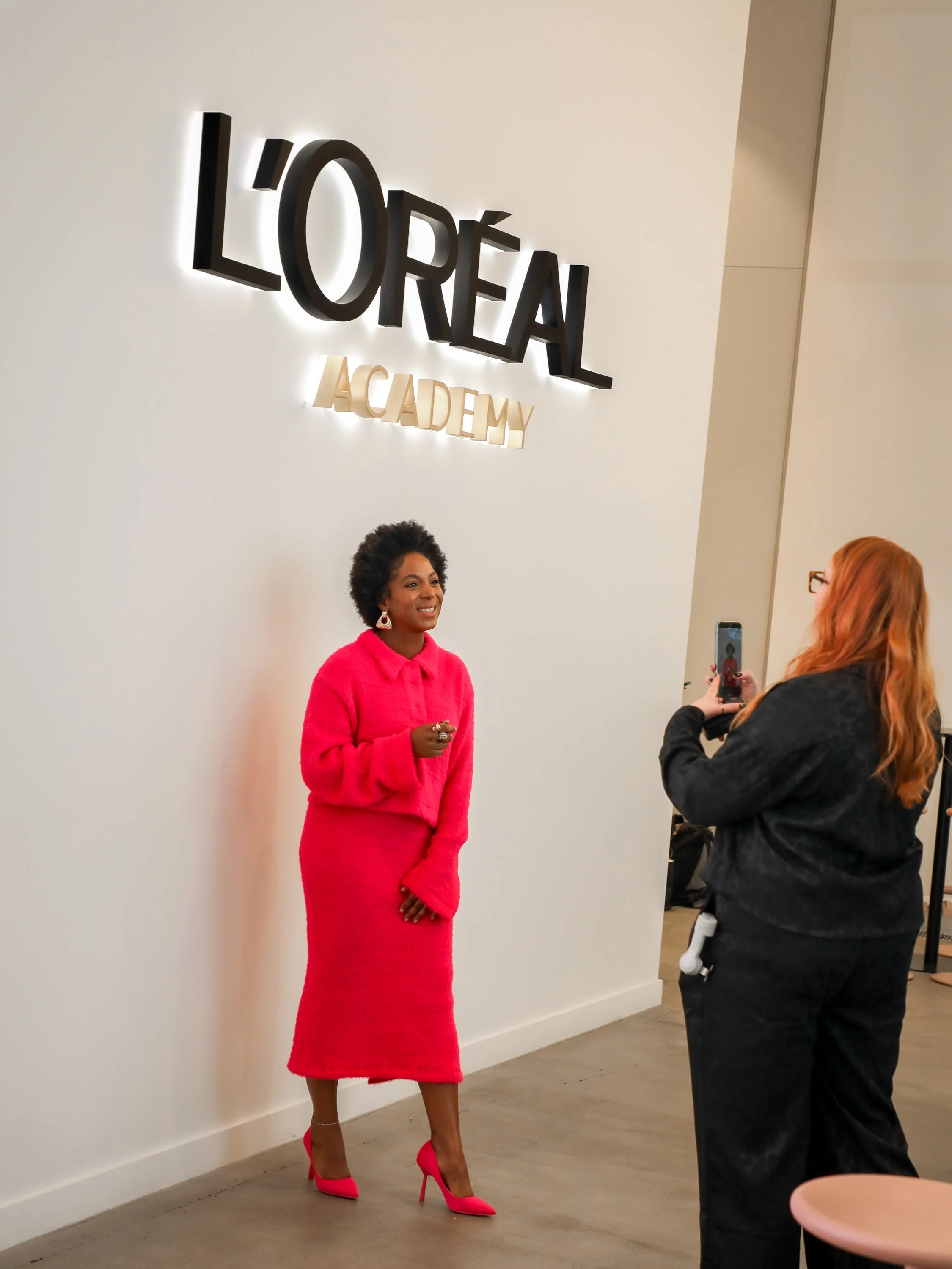 Woman in a bright pink dress and matching high heels standing and smiling in front of a wall with the words "L'Oréal Academy" illuminated above her. Another woman with red hair is taking a photo of her.