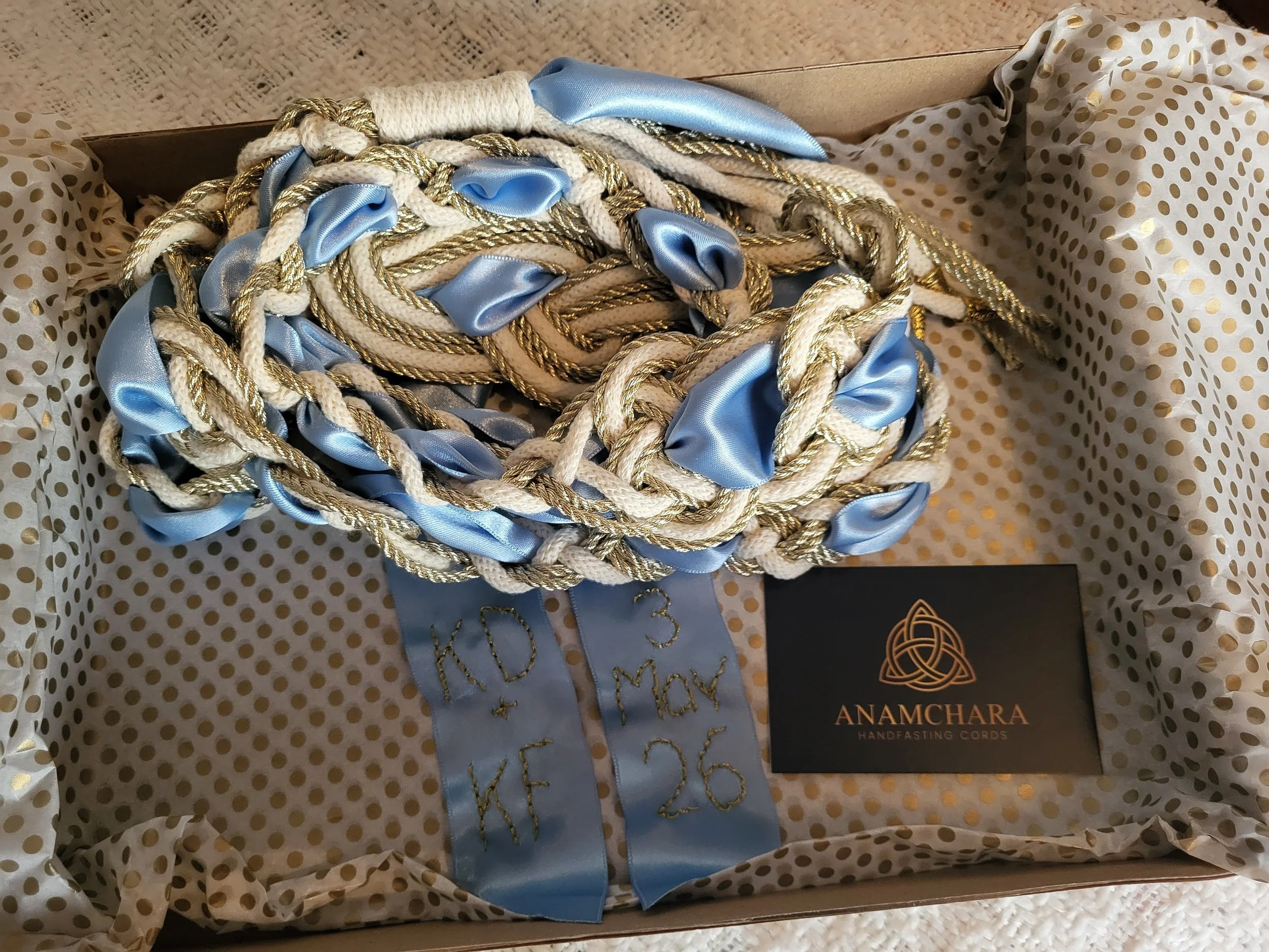 A box of handmade cords with blue satin ribbons, gold and white twisted ropes, and a black card reading 'Anamchara Handfasting Cords'. Wedding tradition, handfasting rope, handtying ceremony, Celtic.