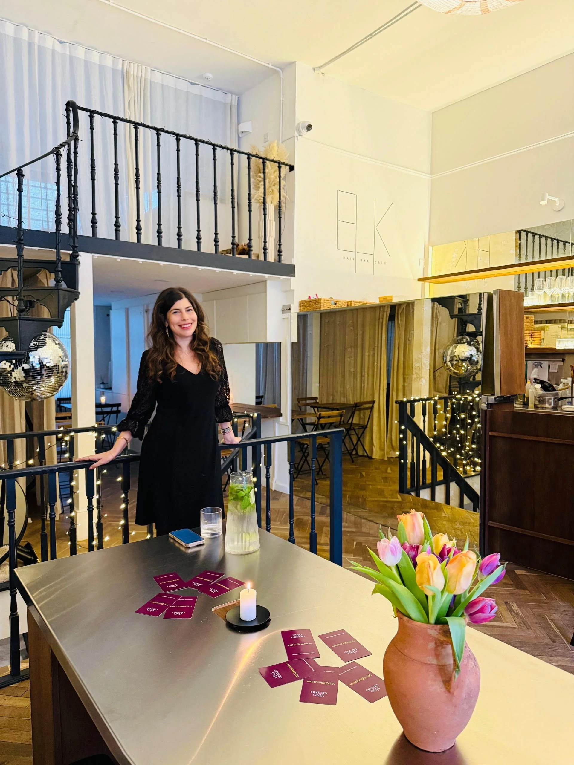 A woman in a black dress with lace sleeves standing behind a table with purple cards, a lit candle, a vase of pink and yellow tulips, and a glass pitcher with mint leaves and lemon. The background shows a cozy interior with string lights, a staircase with disco ball, and a logo on the wall that reads 'HK'.