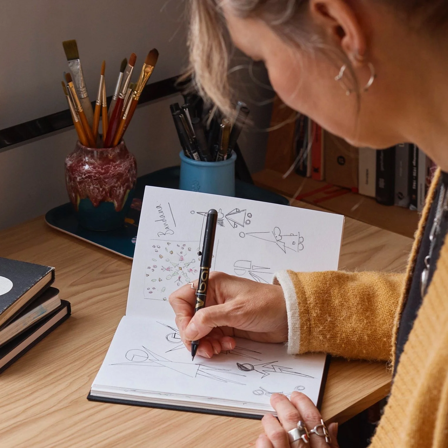 A woman is drawing geometric shapes and diagrams in a notebook with a pen, sitting at a wooden desk. There are two cups with brushes and pens behind her and a closed black notebook on the left side of the desk.