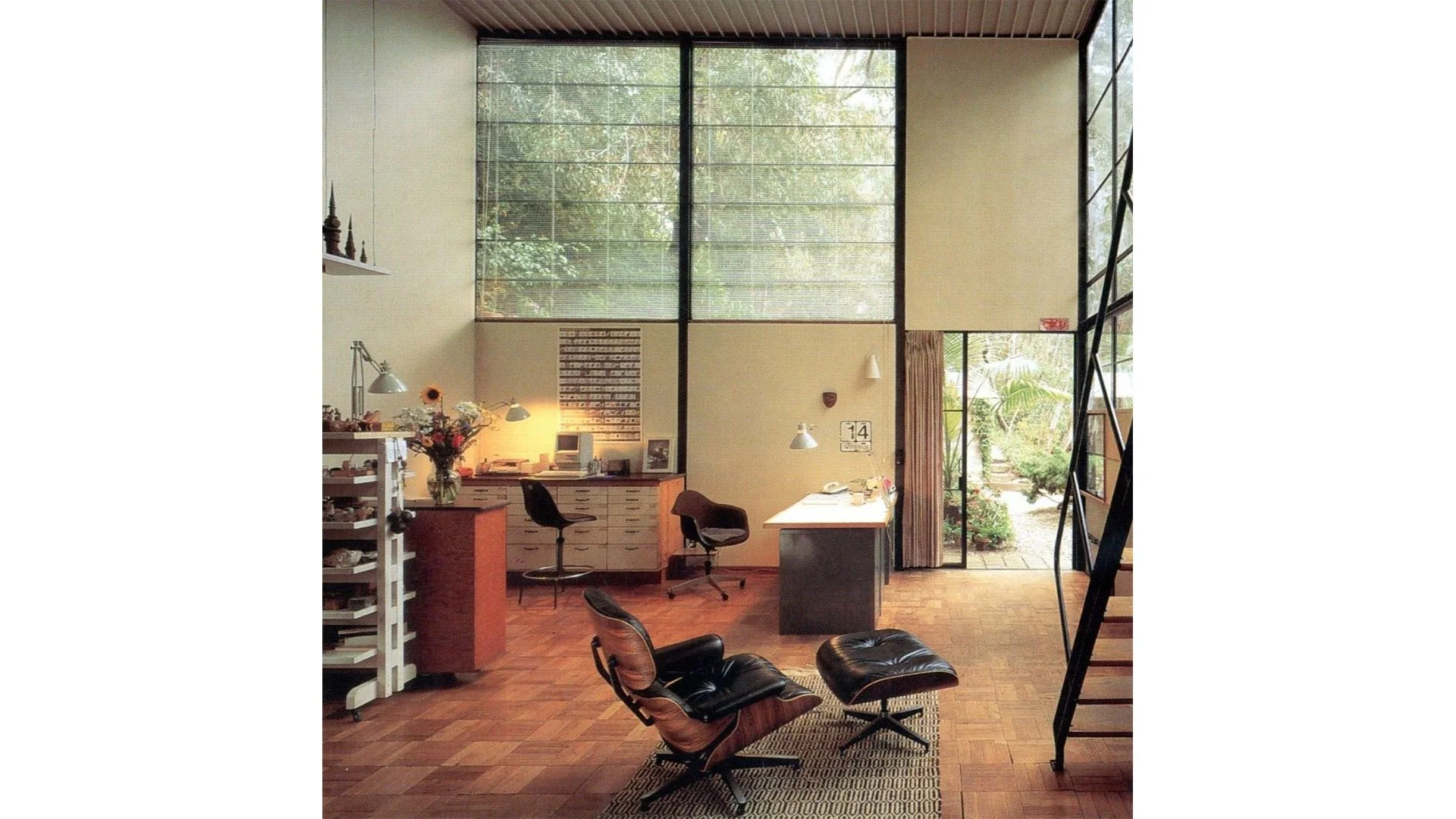 Interior of an office with large windows, corner desk, black chairs, and a lounge chair with ottoman, wooden flooring, and lush greenery visible outside.