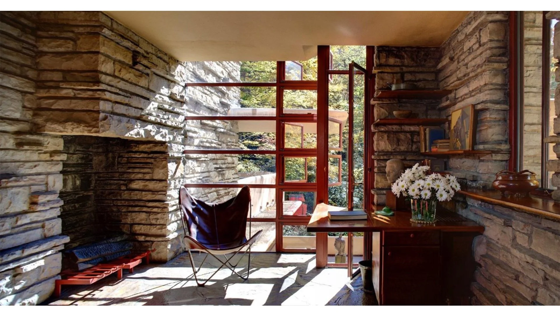 Interior of a room with rustic stone walls, large red-framed windows, a wooden desk with books and a vase of white flowers, a chair, and decorative items like a bust and pottery, with trees visible outside.