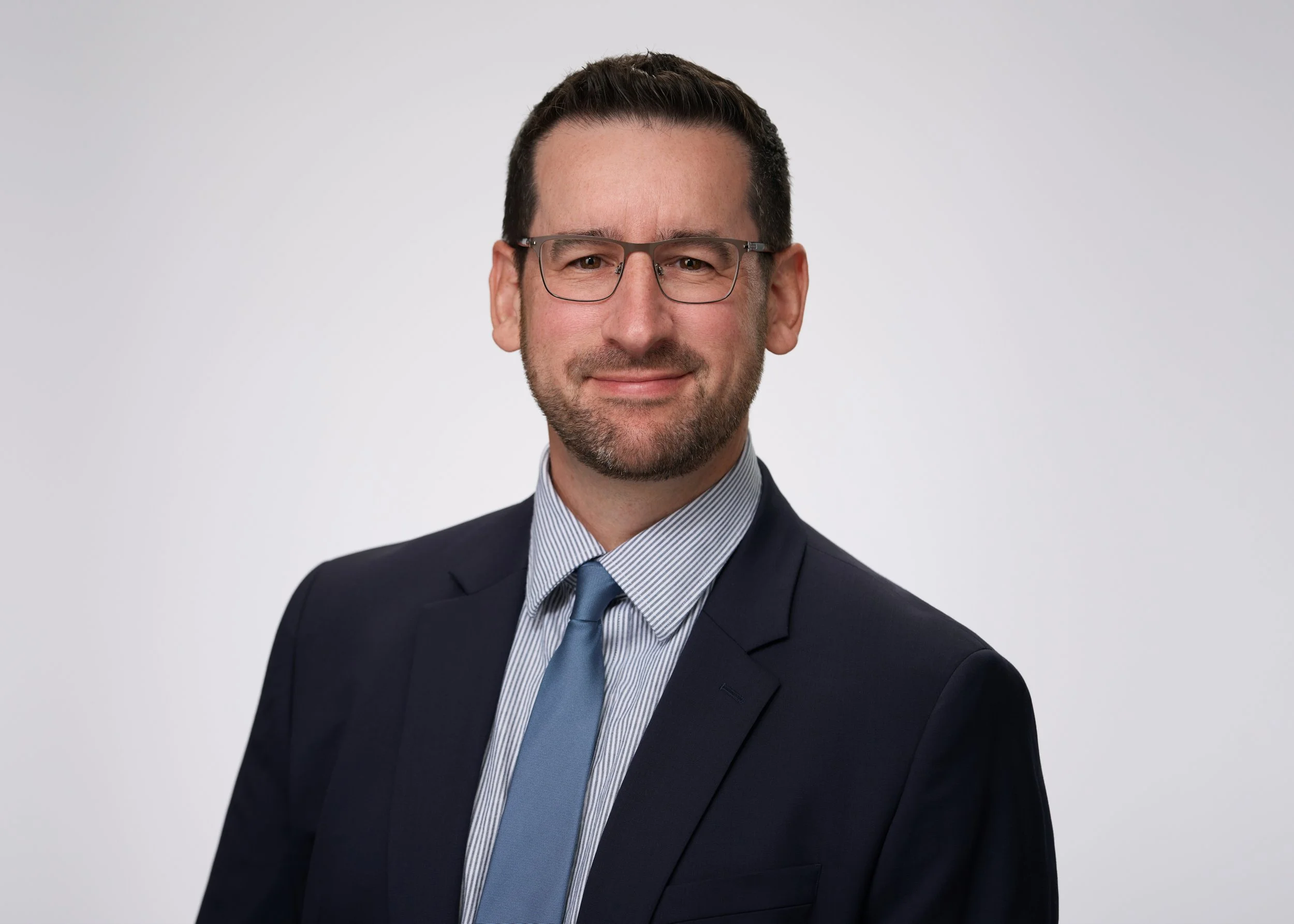 A middle-aged man with glasses, dark hair, and facial hair, dressed in a navy suit, light blue shirt, and matching tie, smiling against a plain gray background.