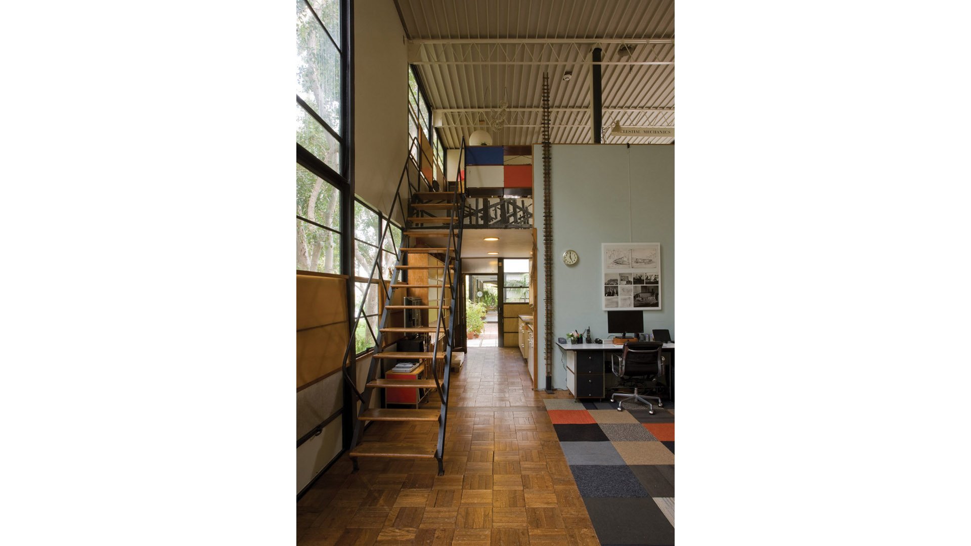 Interior view of a modern office or studio space with high ceiling, large windows on the left, a staircase with wooden steps and metal railing leading to a mezzanine, a desk with a computer on the right, and a colorful rug on the wooden floor.
