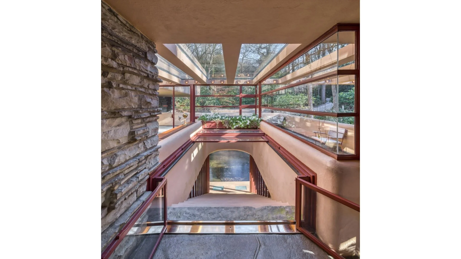 Interior view of a modern home with a glass enclosed balcony area, visible staircase leading down, decorative stone wall on the left, and large windows overlooking a garden with trees.
