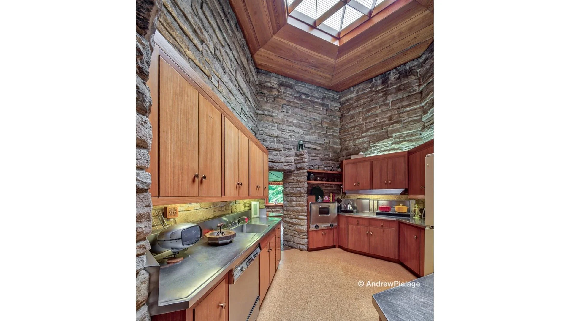 Kitchen with stone walls, wooden cabinets, and a sloped wooden ceiling with a skylight. Countertop has kitchen appliances and utensils.