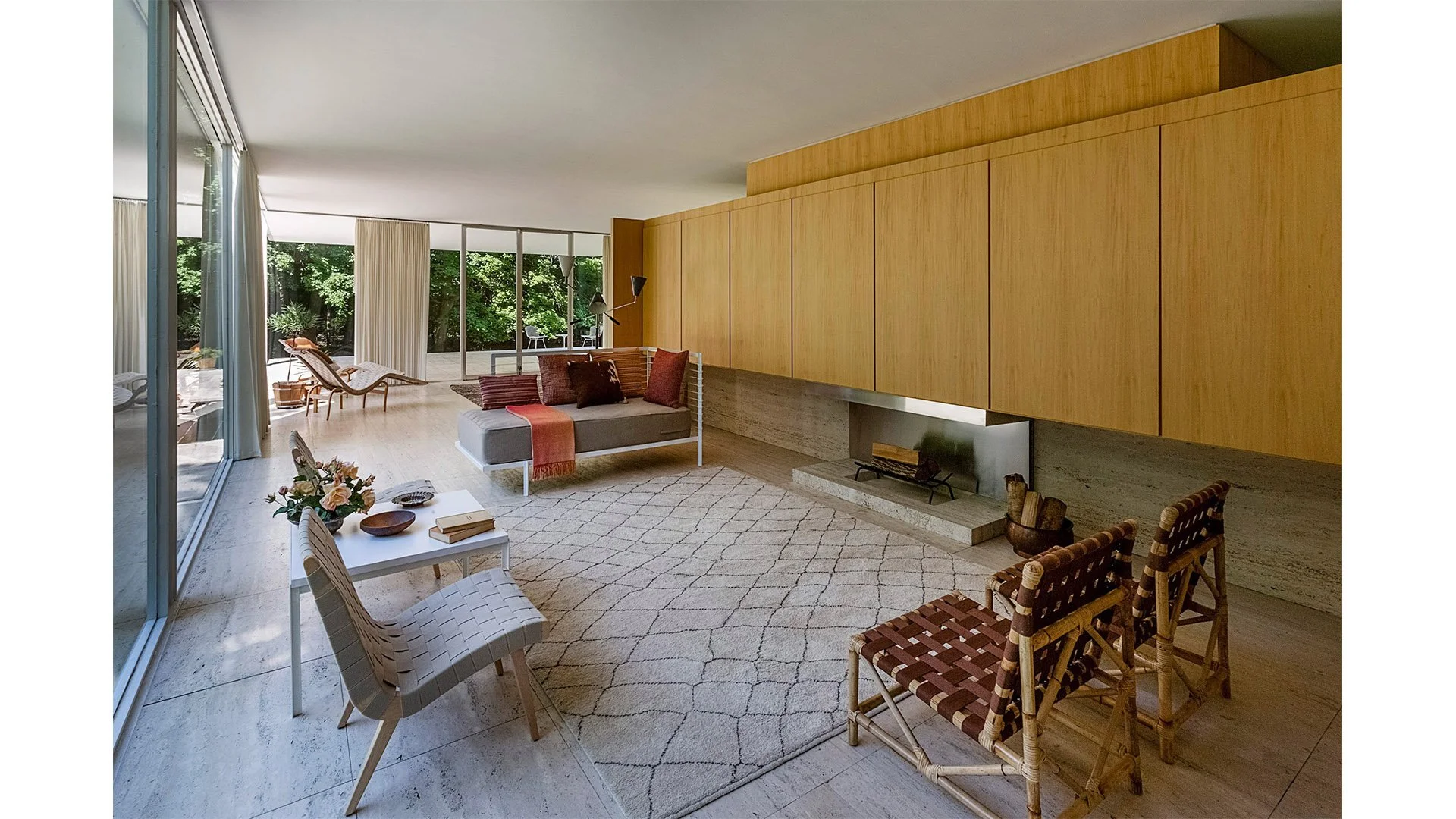Living room with large sliding glass doors leading to outdoor space, a light-colored area rug, a low white coffee table with books and flowers, a modern gray sofa with brown and rust pillows, a built-in wood wall unit, and two rattan chairs.