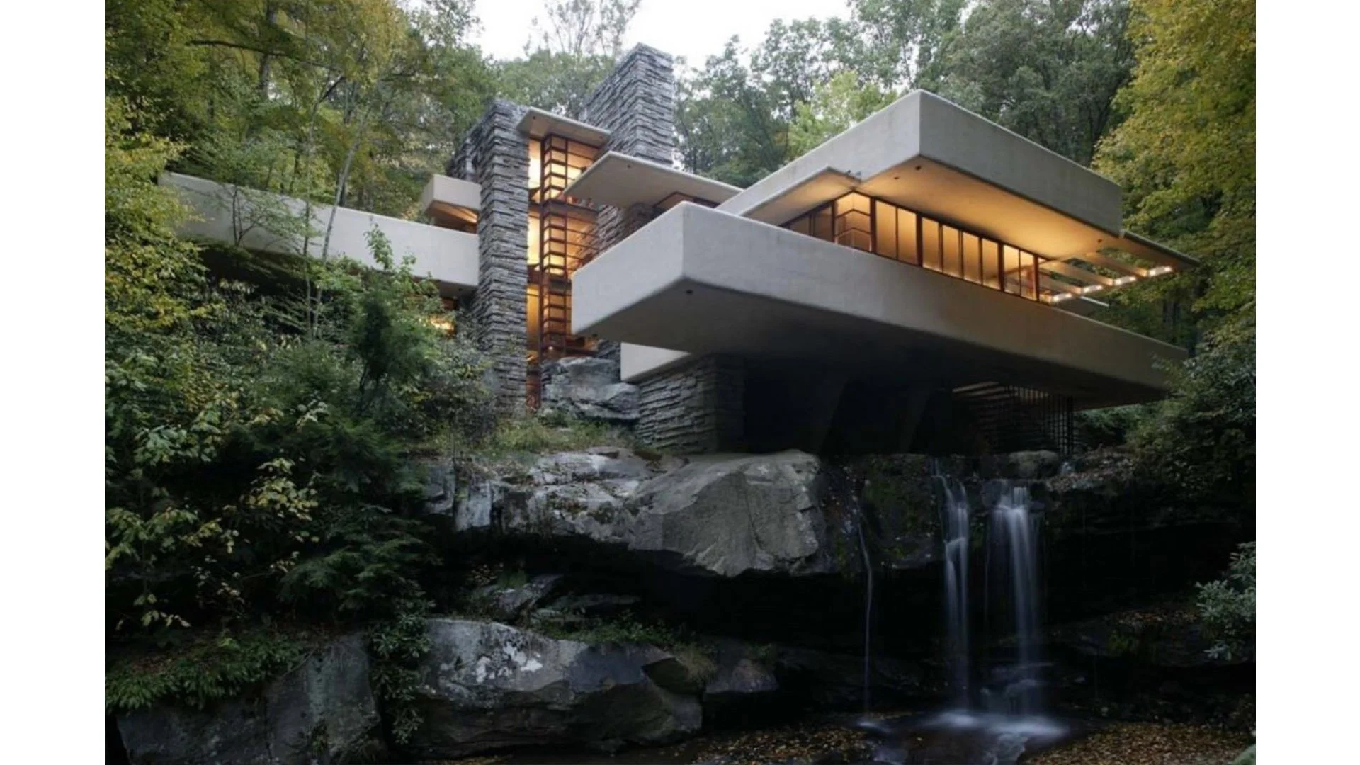 Modern house built into a hillside with large concrete balconies, stone walls, and large windows surrounded by trees and a waterfall feature.