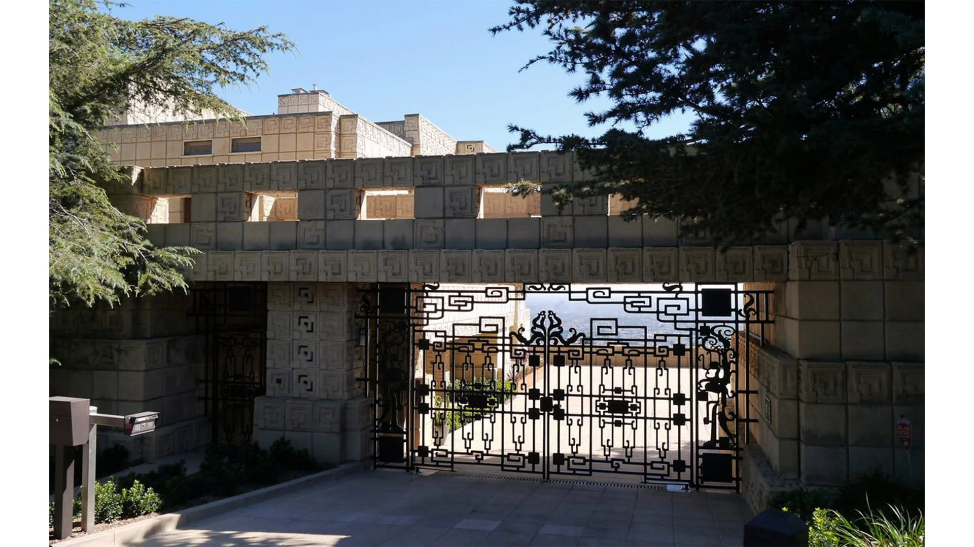 Image of a decorative stone wall and an ornate metal gate, with trees on either side and a clear blue sky in the background.