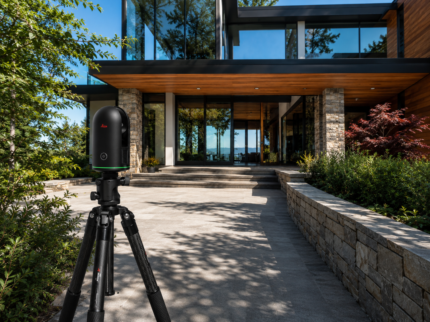 A laser scanner on a tripod in front of a modern house with glass windows, surrounded by greenery and trees, on a sunny day.