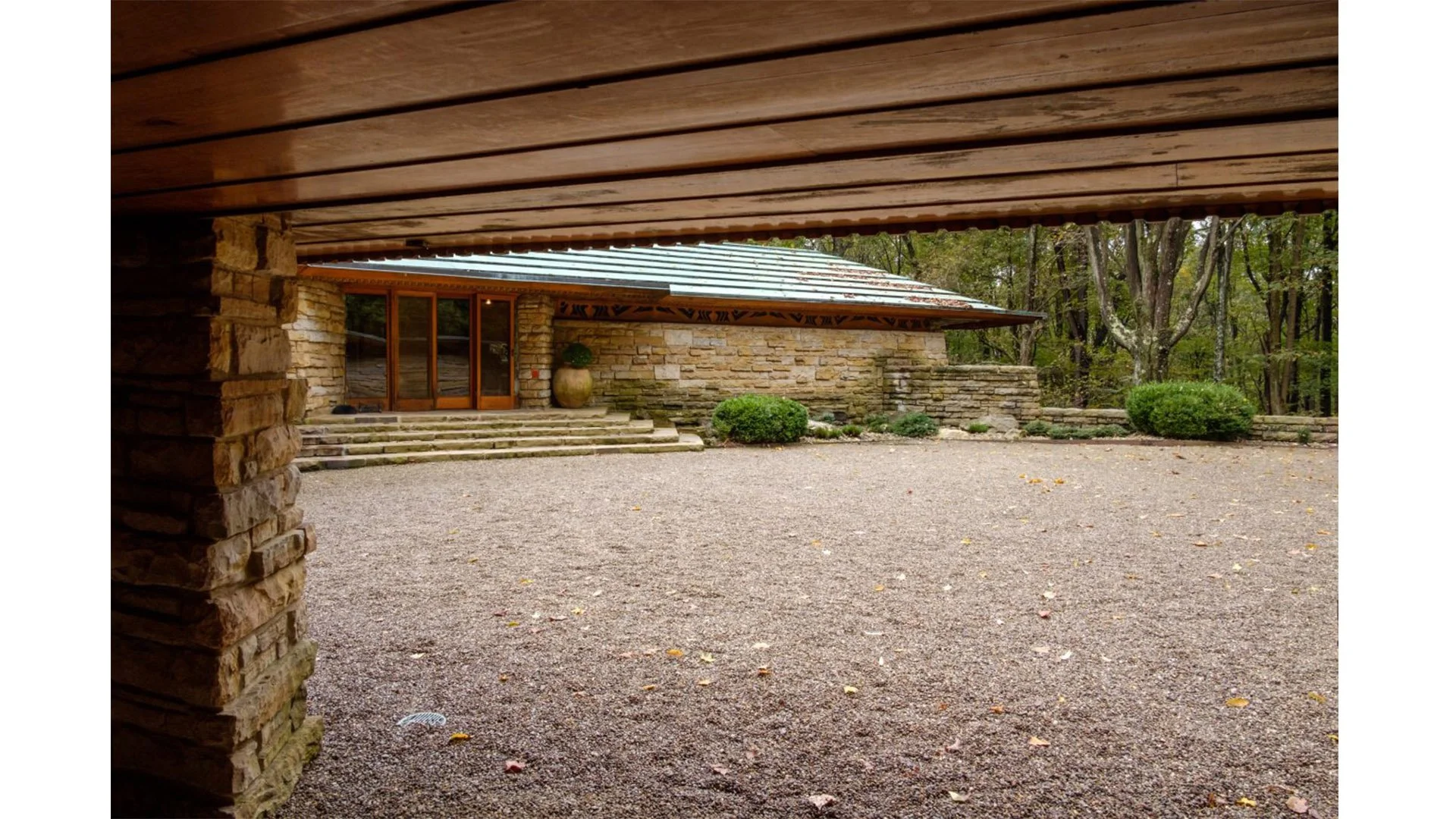 View of a house with stone walls and a metal roof, seen from under a porch, with steps leading up to a wooden door, surrounded by trees and bushes.