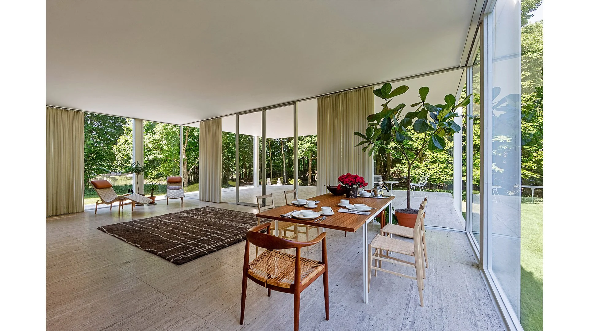Modern open living space with large glass windows showing green outdoor scenery, a wooden dining table with white dishes, a tall potted plant, and lounge chairs near the windows.