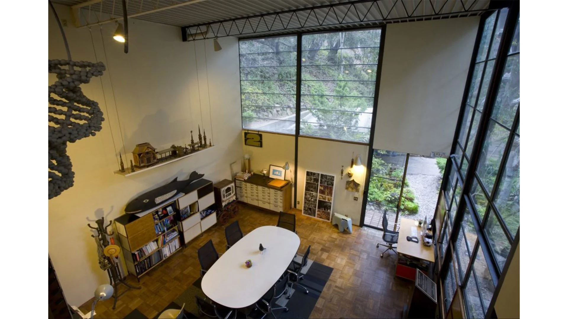 A high-angle view of a modern office with large windows, a white oval conference table surrounded by black chairs, bookshelves, and decorative items.