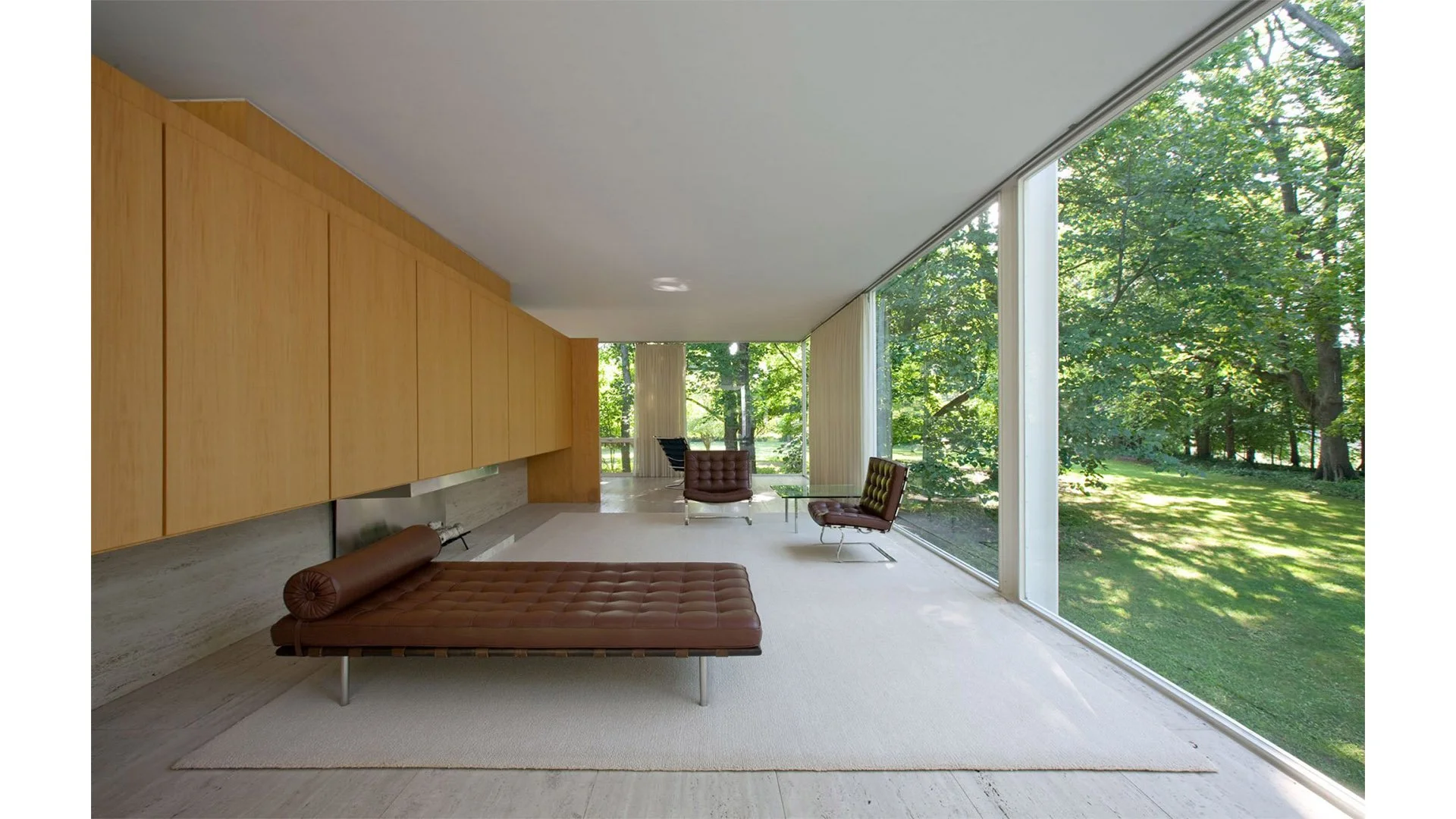 Minimalist living room with large floor-to-ceiling windows showing a lush green yard, wooden cabinets on the left, a brown leather daybed on a white rug, and two brown chairs with metal frames.