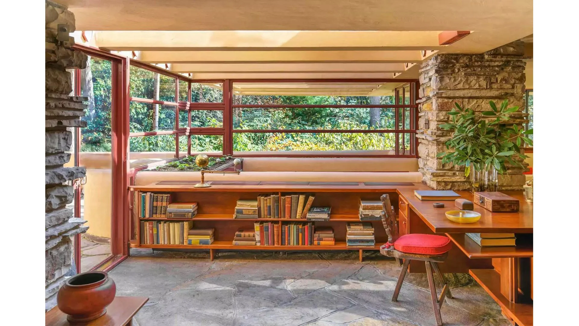 A cozy corner with stone and wood design, large windows revealing greenery outside, a wooden bookshelf filled with books, and a wooden desk with a potted plant, a yellow dish, and a red cushioned stool.